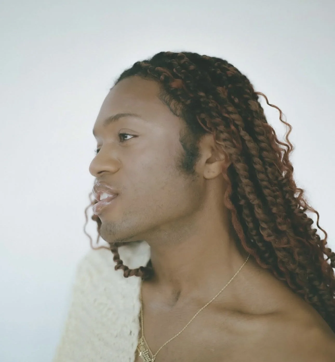 A young person with dark skin and long, reddish-brown, curly dreadlocks looks to the side against a plain white background.