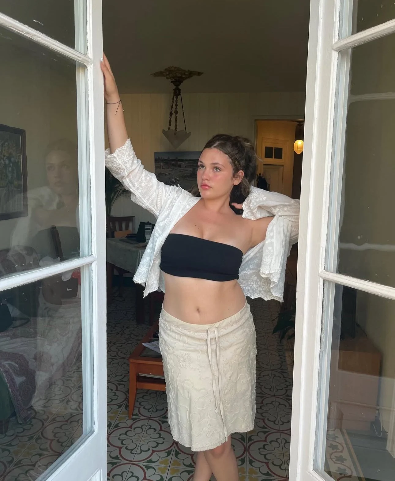 A young woman with dark hair in a messy bun, wearing a black tube top, a cream-colored skirt, and an open white lace jacket, standing in a doorway and looking off to the side with one arm raised and the other resting behind her head.