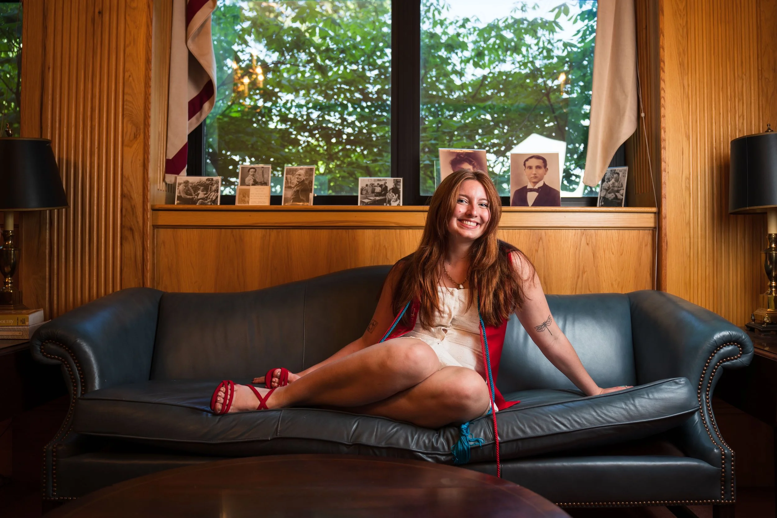 A woman with long reddish-brown hair sitting on a dark leather couch in a wooden-paneled room with framed photographs on a windowsill behind her, smiling at the camera.
