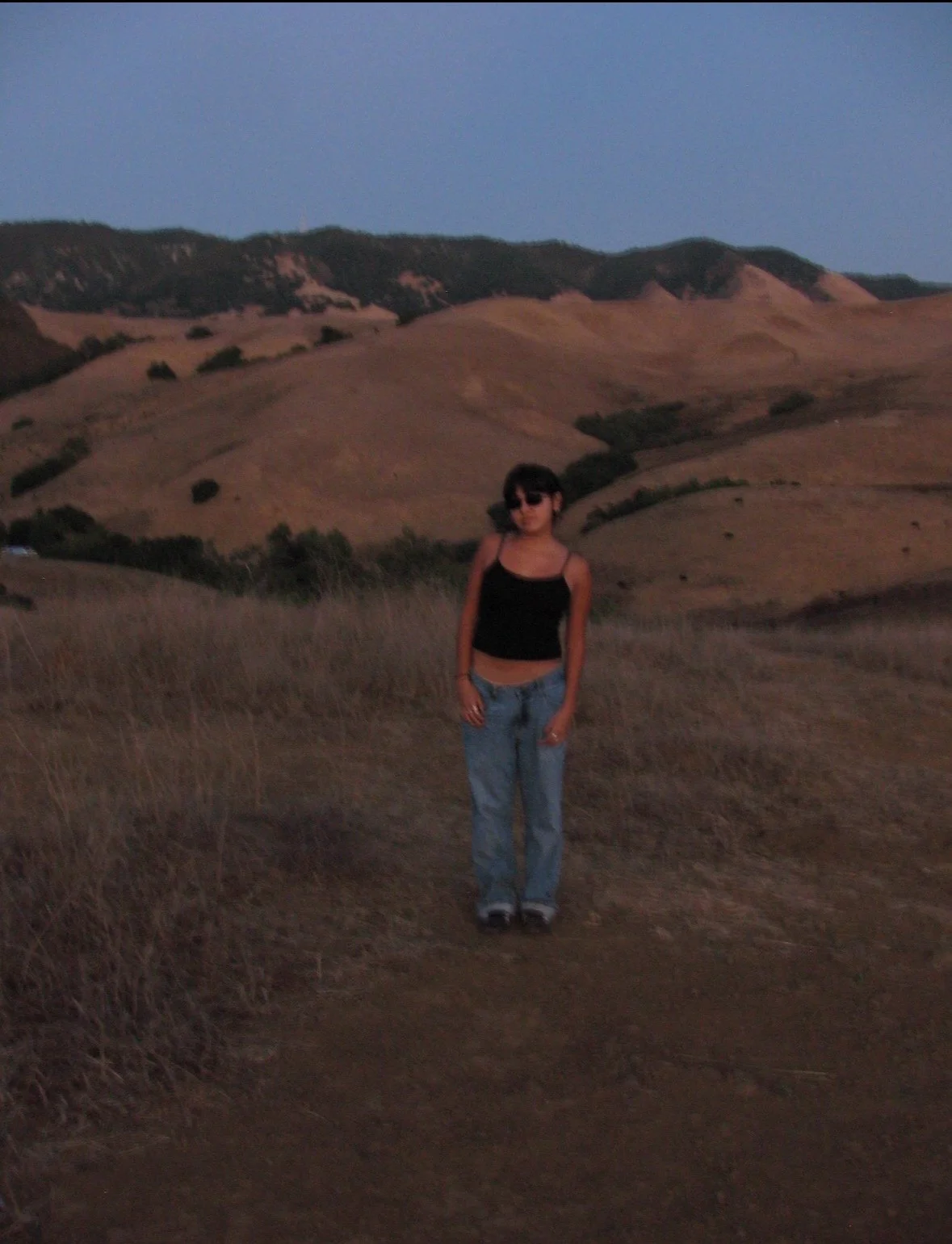 A young woman with dark hair and sunglasses stands in a dry grassy field with rolling hills in the background during dusk.