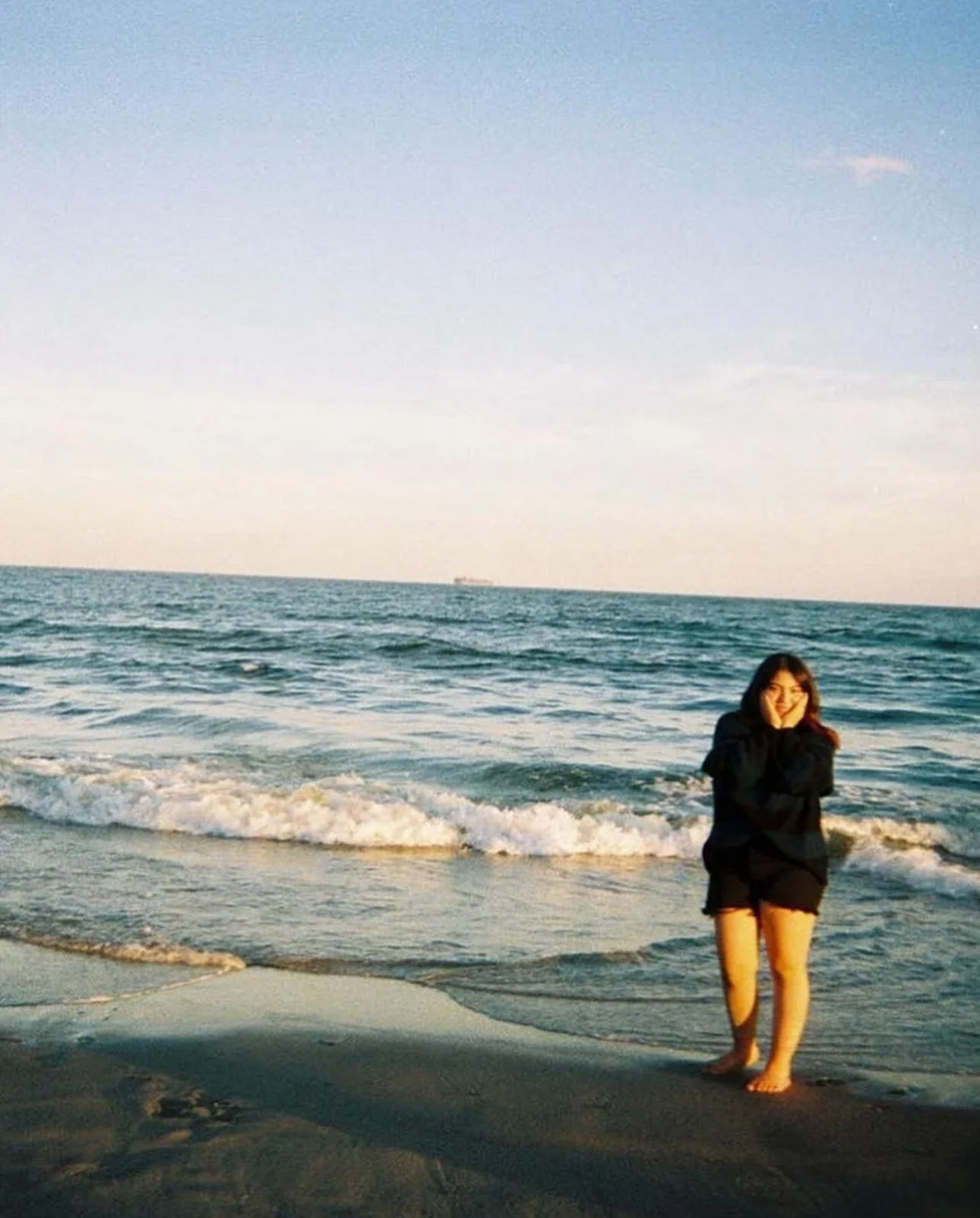 A girl in black jacket and shorts standing barefoot on the sand at the beach, smiling with hands around her face, with waves and a ship in the background.