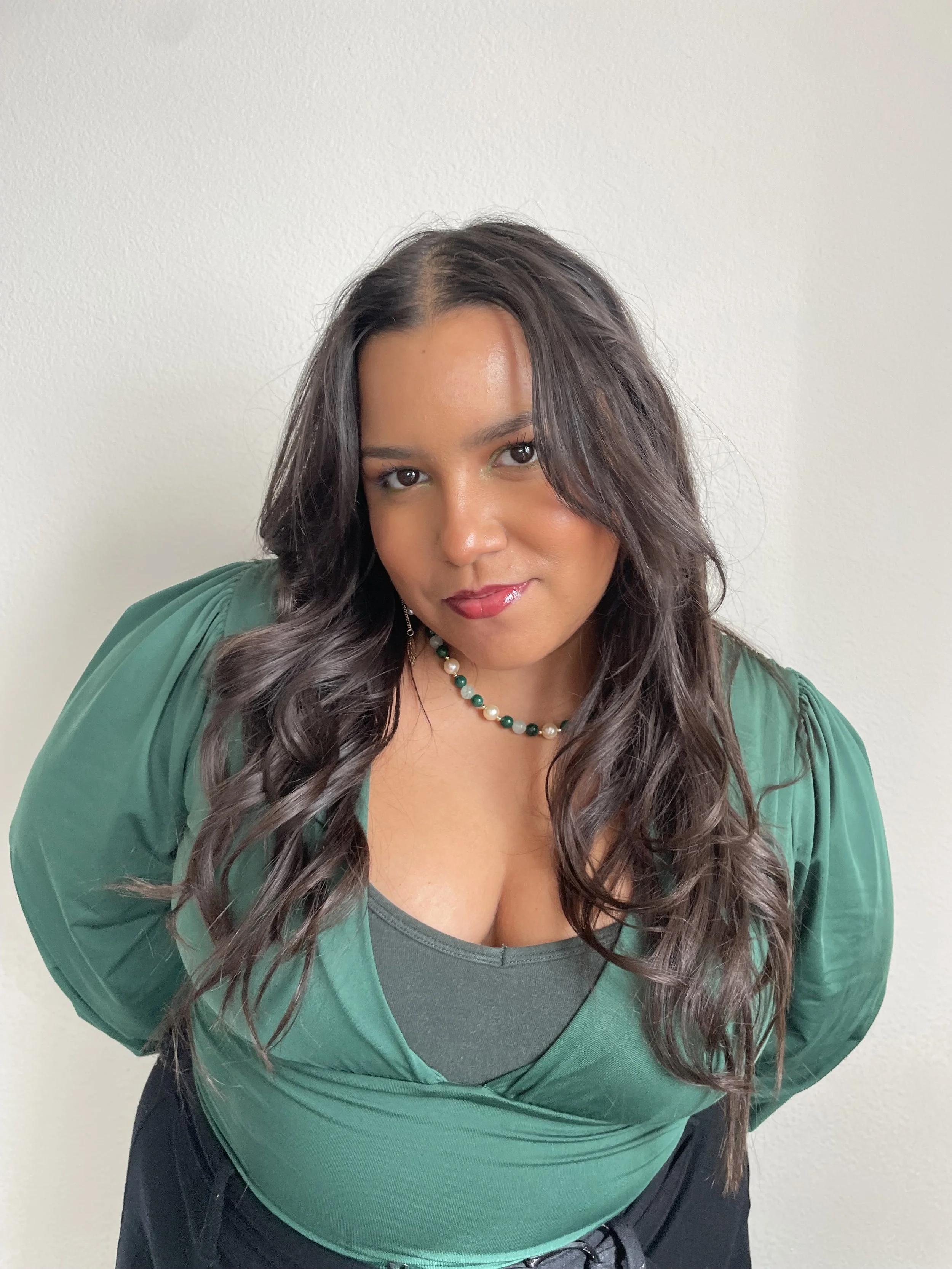 A woman with long, wavy dark hair, wearing a green top with gathered sleeves, a black necklace with green and white beads, and red lipstick, is leaning slightly forward and looking at the camera against a plain white wall.
