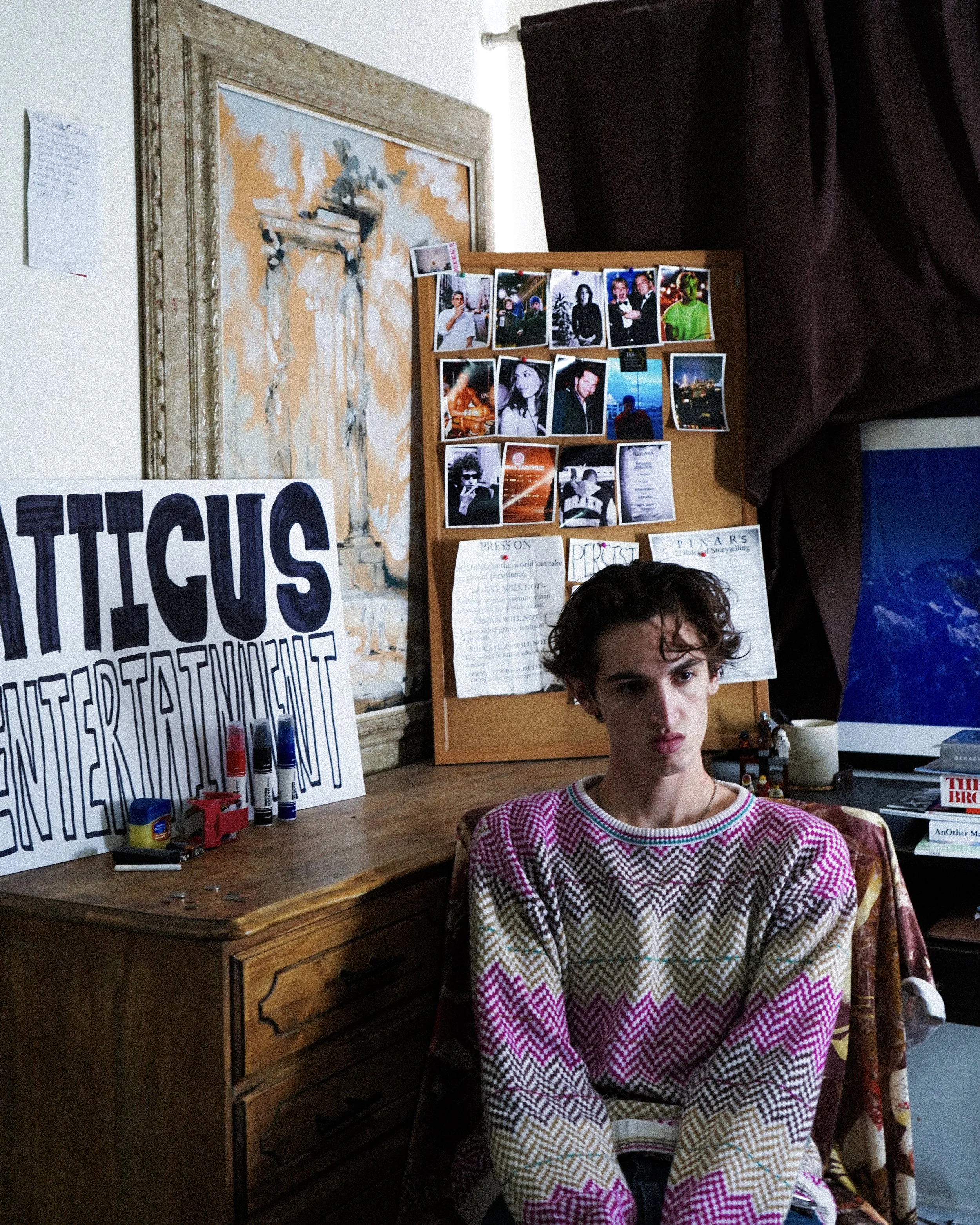 Young person sitting in a room with a wooden desk, bulletin board, and various posters and photos on the wall. They are wearing a colorful patterned sweater.