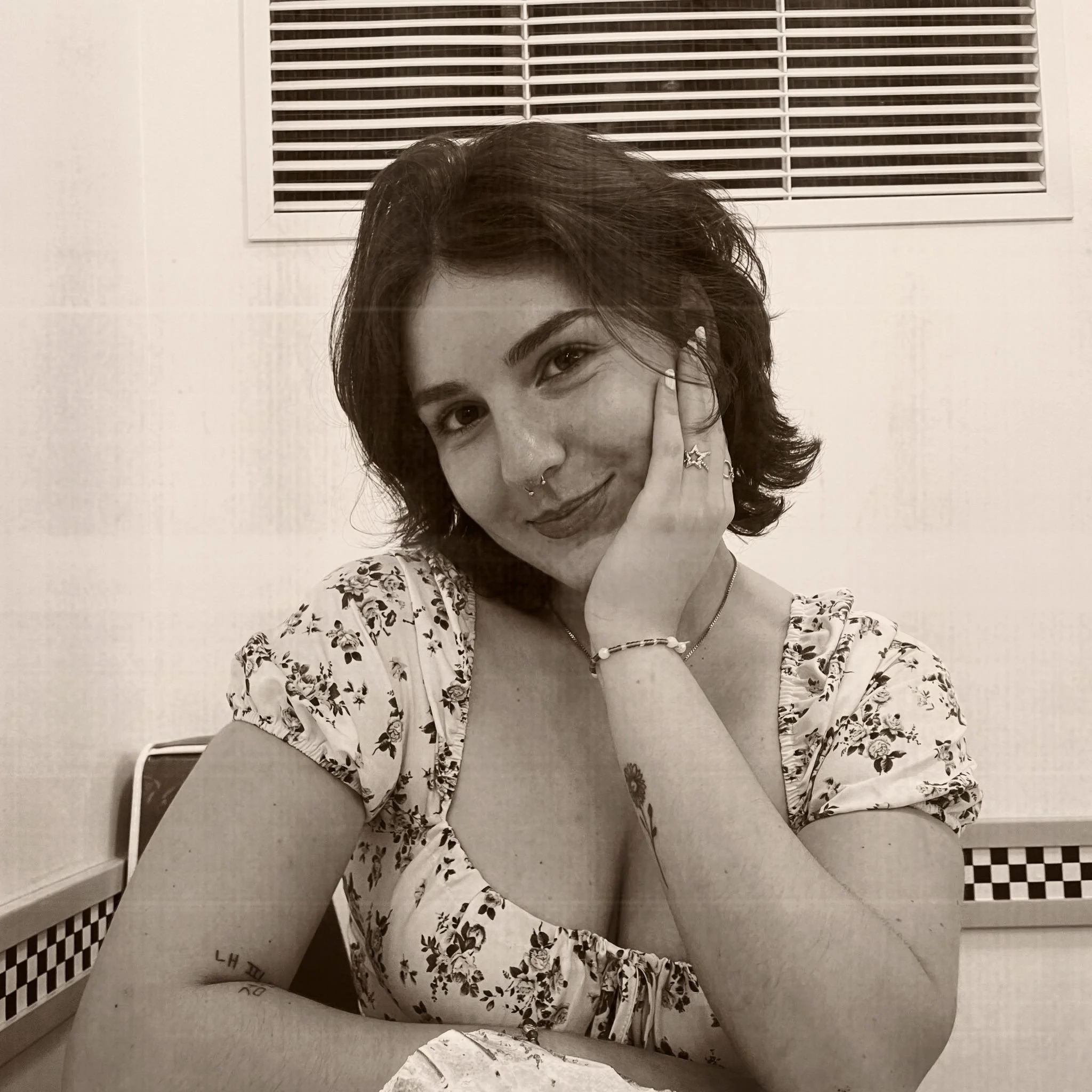 A young woman with short, wavy hair and tattoos on her arms sitting at a table, smiling at the camera, with her hand resting on her face. She is wearing a floral dress, a necklace, rings, and a bracelet.
