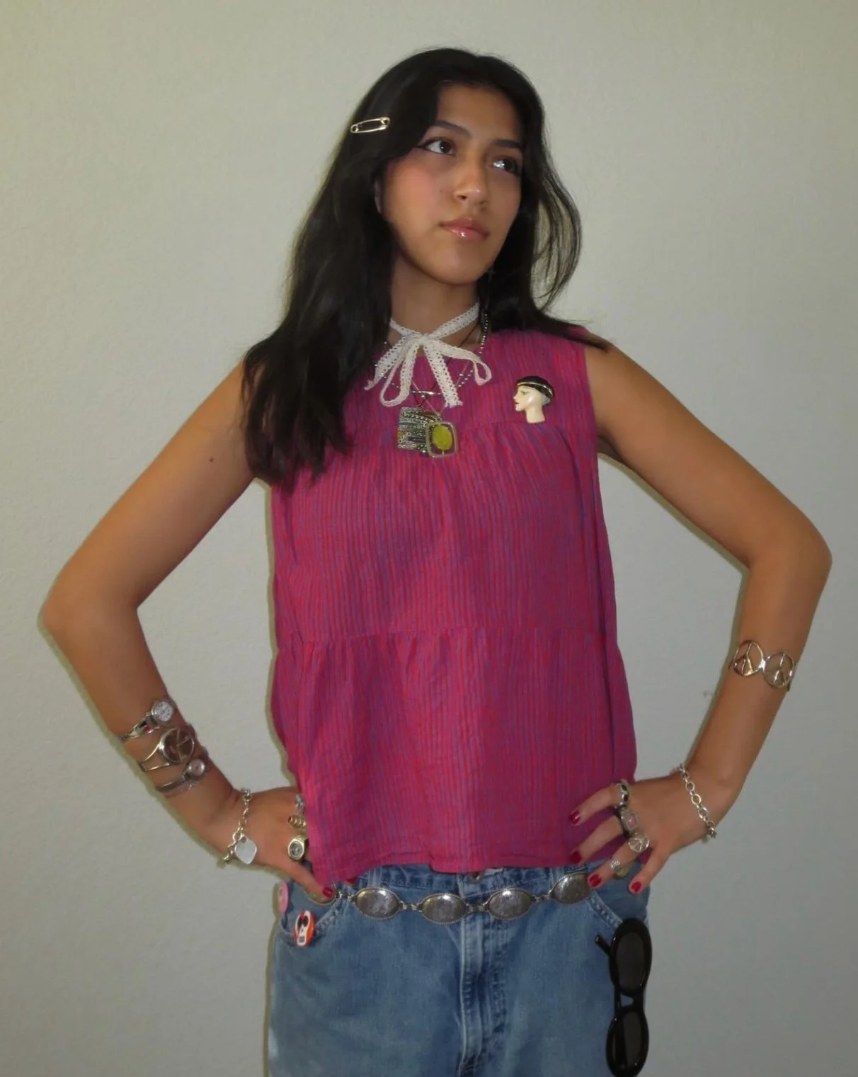 Young woman with long dark hair wearing a pink sleeveless top and blue jeans, accessorized with jewelry, standing with hands on hips against a plain wall.