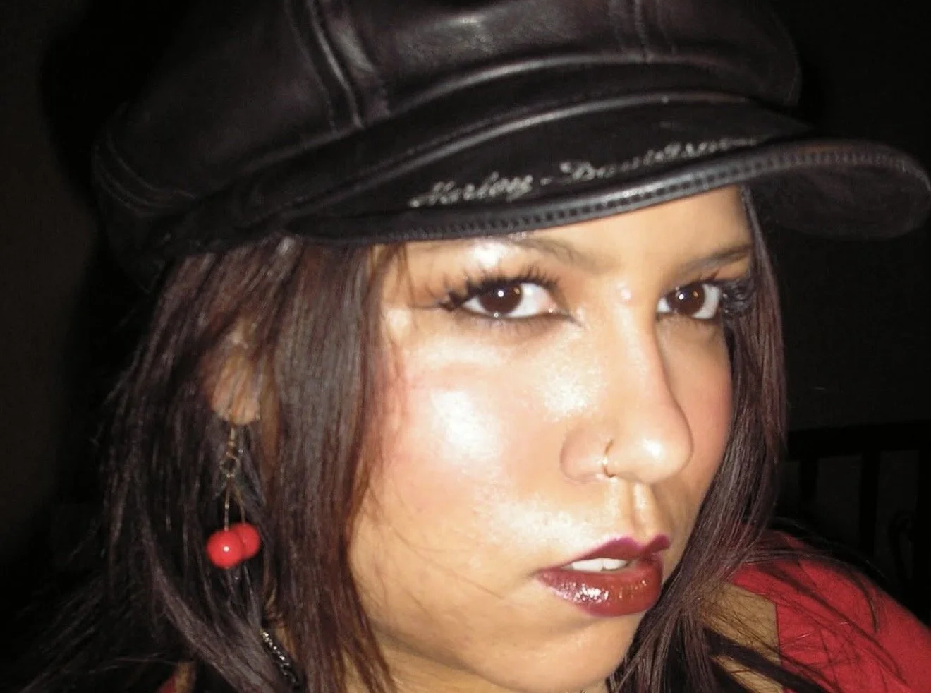 A woman with long, wavy dark hair, wearing a green top with gathered sleeves, a black necklace with green and white beads, and red lipstick, is leaning slightly forward and looking at the camera against a plain white wall.