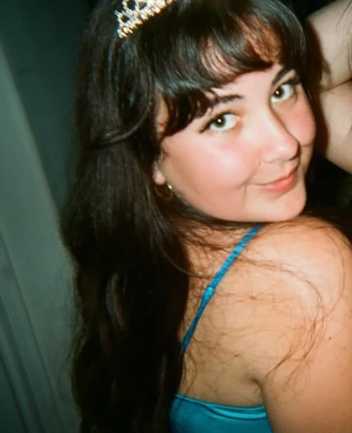 Young woman with dark hair wearing a tiara and a blue spaghetti strap top, smiling at the camera.