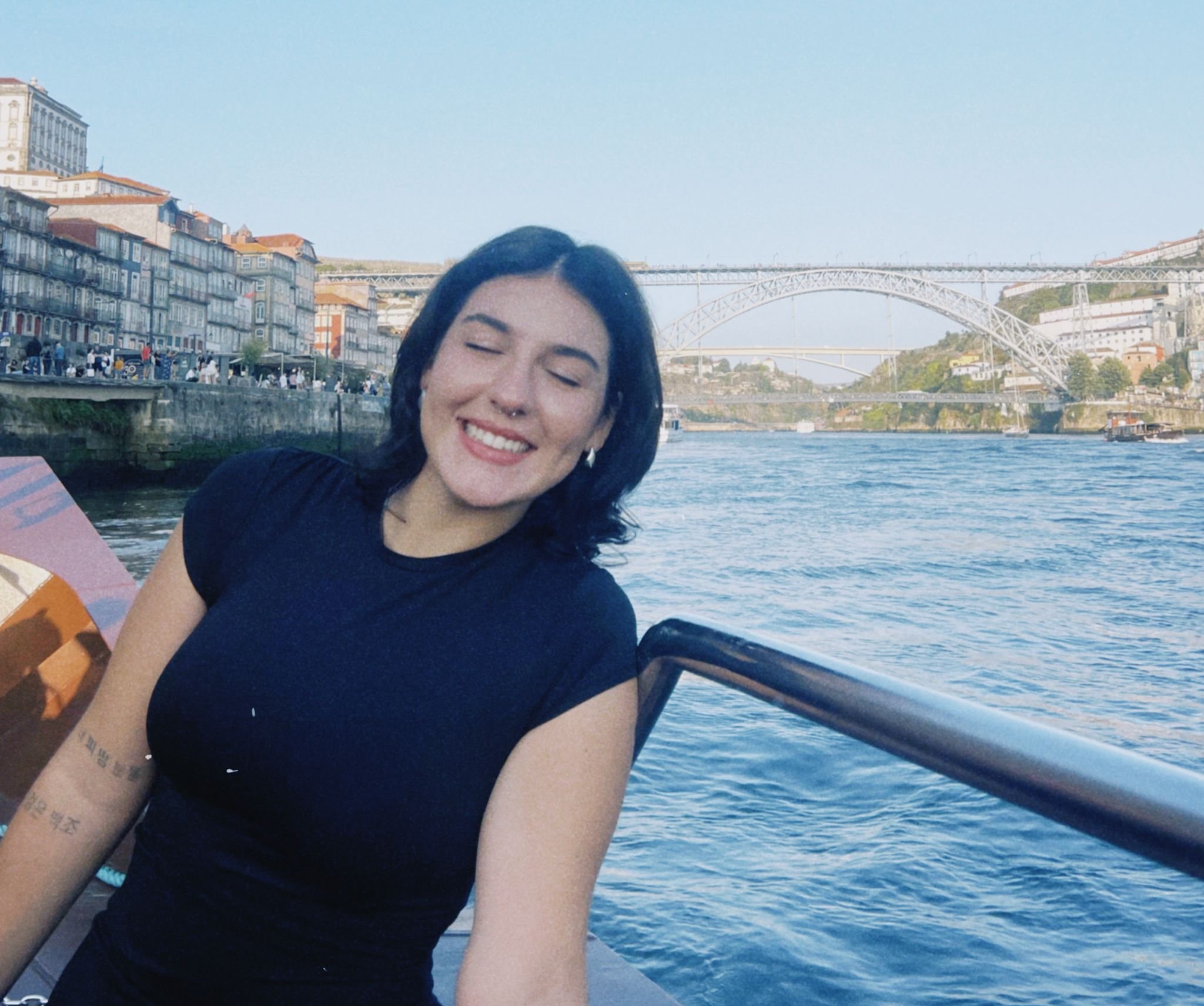 A young woman with short, wavy hair and tattoos on her arms sitting at a table, smiling at the camera, with her hand resting on her face. She is wearing a floral dress, a necklace, rings, and a bracelet.