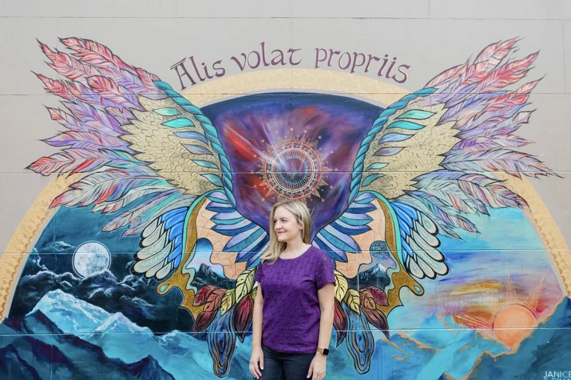 A woman with blonde hair wearing a purple top and black pants standing in front of a colorful mural of a butterfly with cosmic and mountain scenes inside its wings and the phrase 'Alis volat propriis' written above.