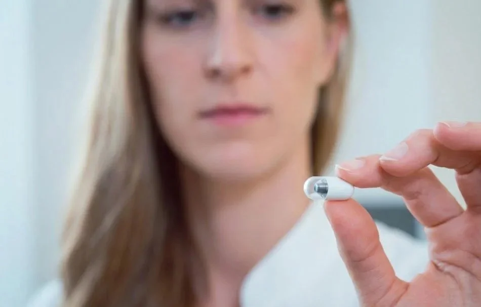 A woman with long brown hair holding a small white and silver hearing aid.