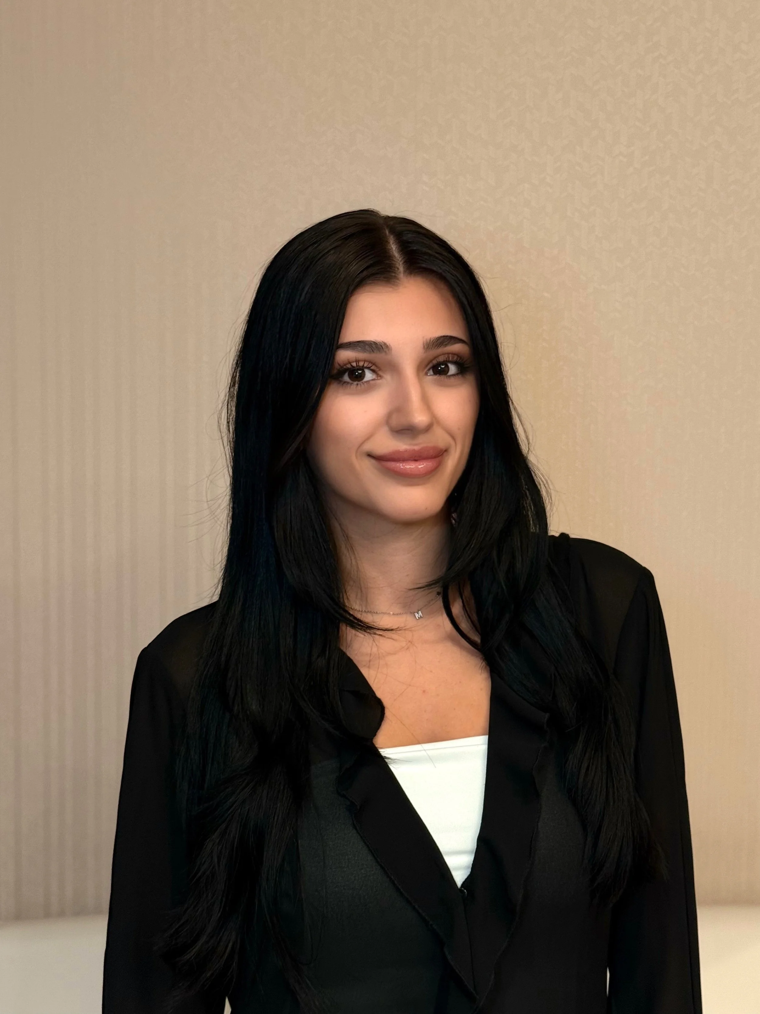 A young woman with long black hair and brown eyes, wearing a black blazer over a white top, standing in front of a plain beige wall.