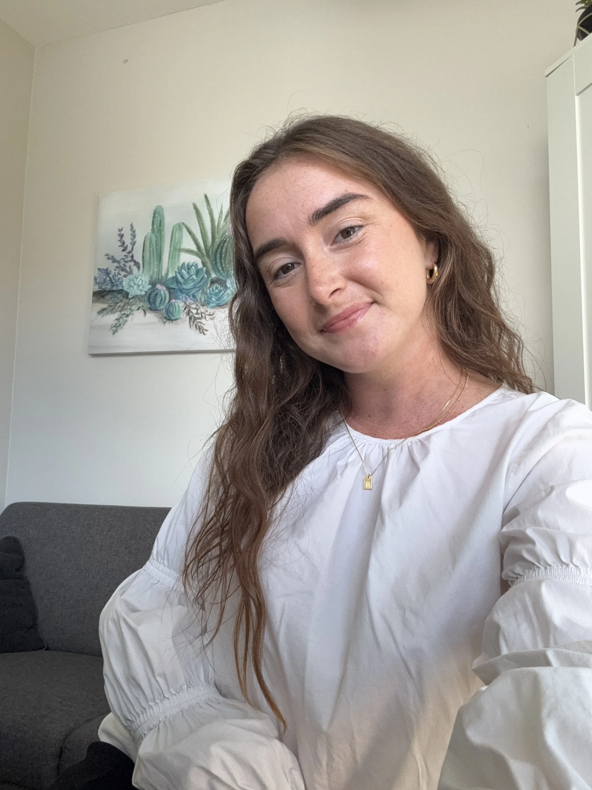 A woman with long wavy brown hair taking a selfie indoors, wearing a white blouse and jewelry, with a painting of cacti and succulents on the wall behind her.