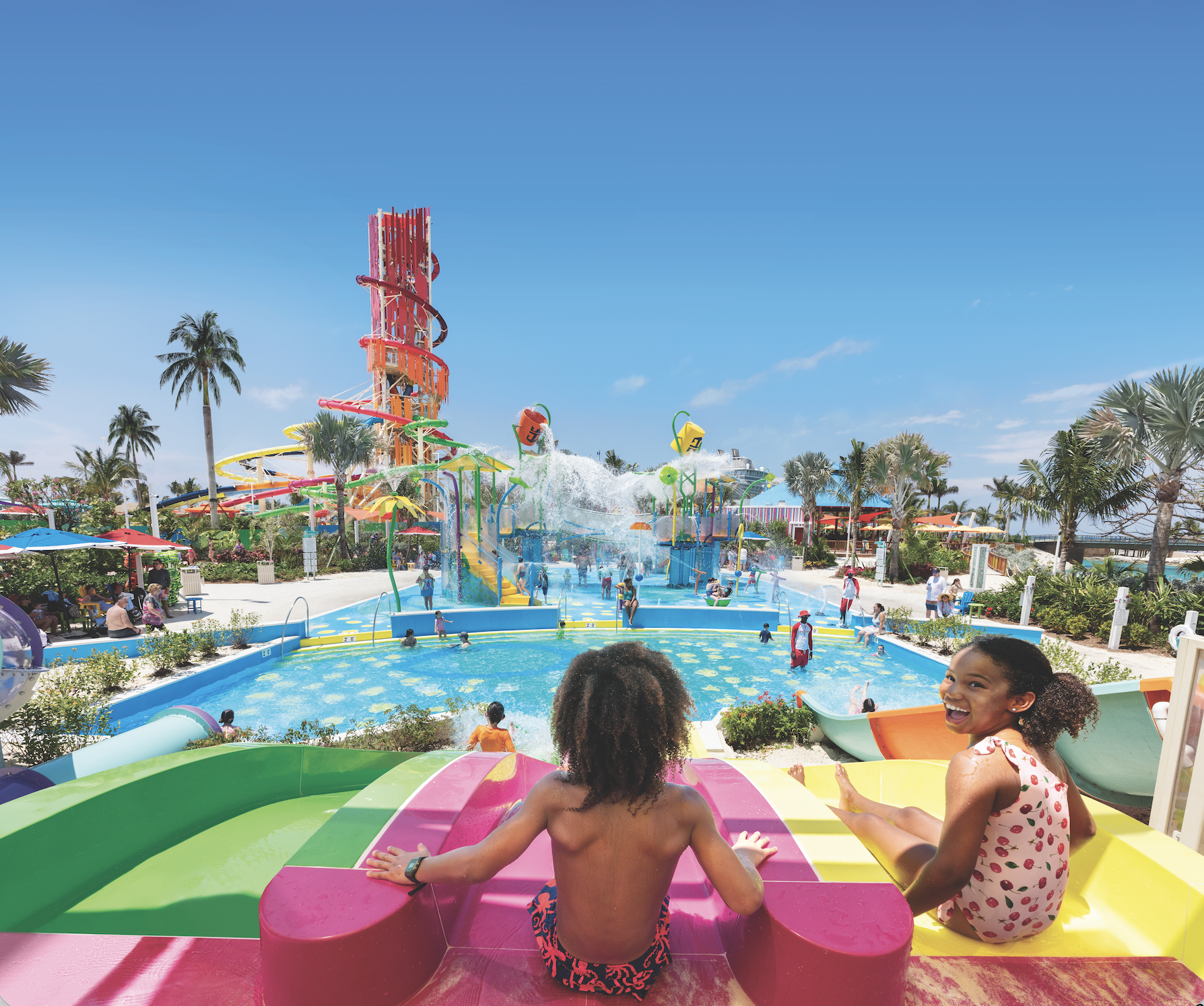 Children getting ready to race down colorful waterslides at Splashaway Bay on Perfect Day at CocoCay, with water play areas, pools, palm trees, and the towering Daredevil’s Peak slide in the background under a sunny Bahamian sky.
