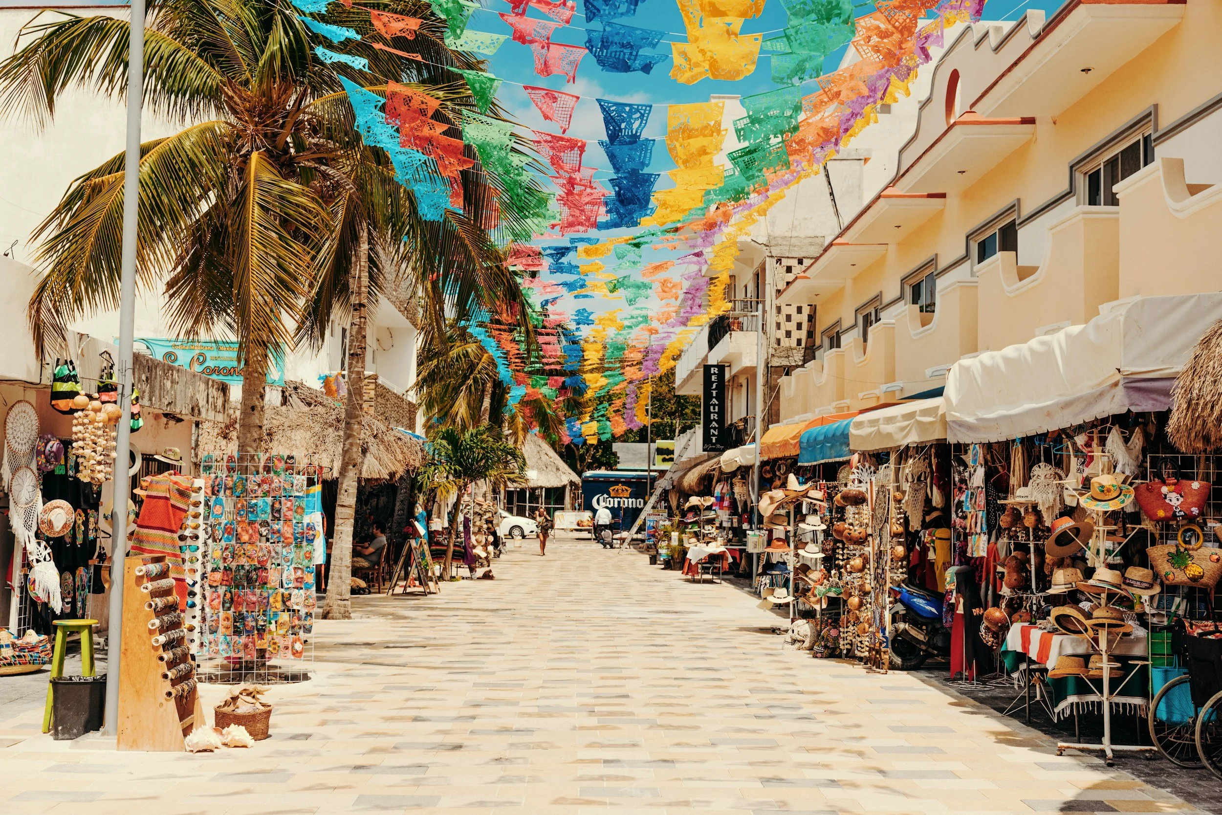 Colorful shopping street in Cozumel lined with local market stalls, souvenirs, palm trees, and vibrant papel picado banners overhead.