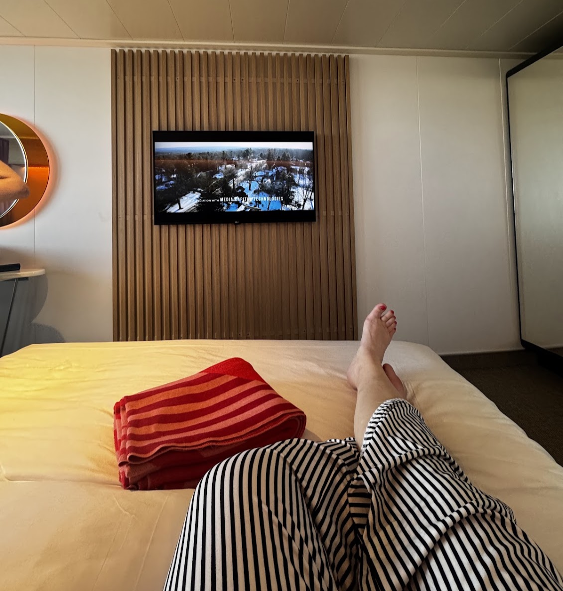 View from the bed inside a modern cruise ship cabin with a wall-mounted TV, wood accent wall, and contemporary design.