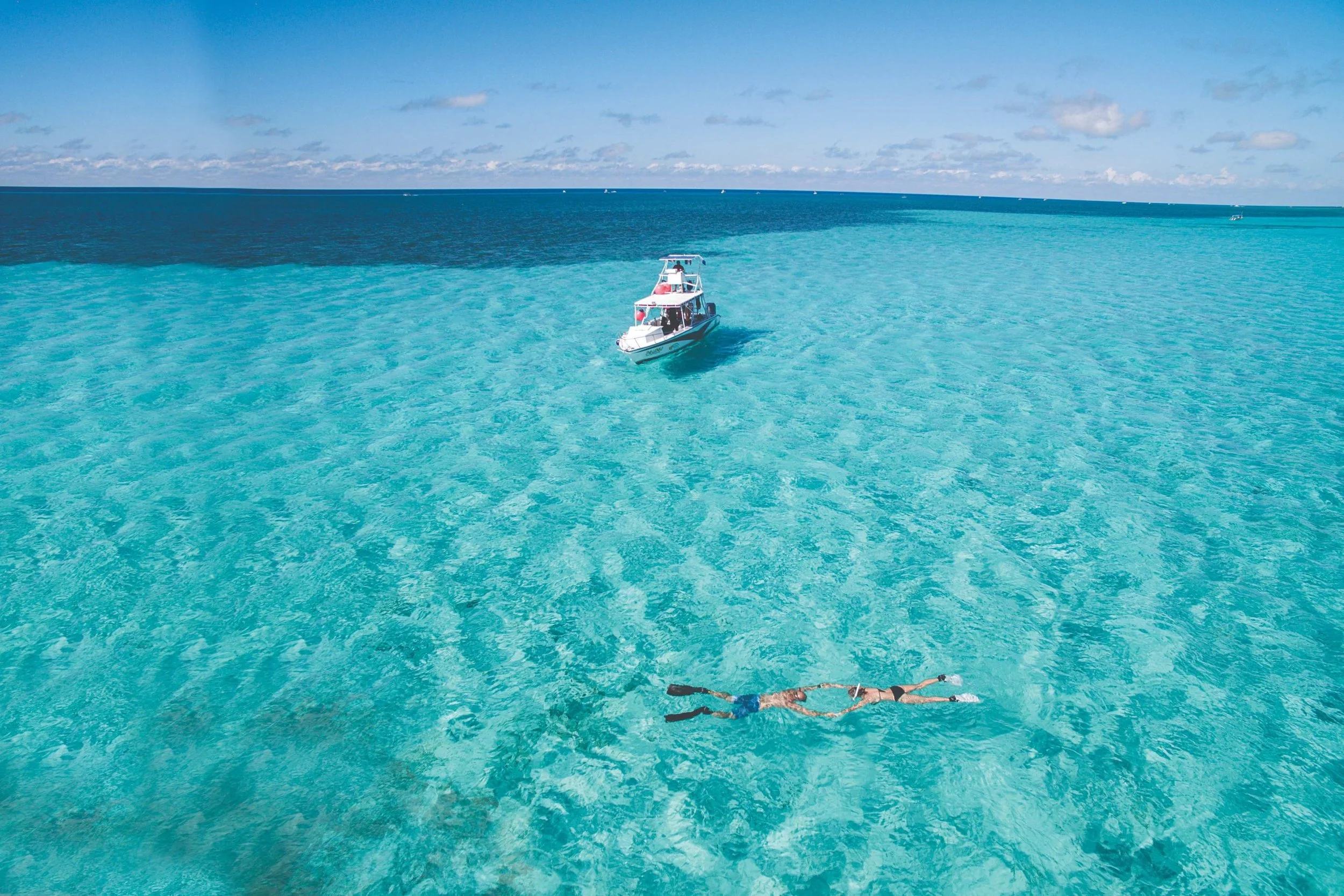Aerial view of two snorkelers floating in crystal-clear turquoise water near a small boat in the Caribbean Sea off the coast of Cozumel.