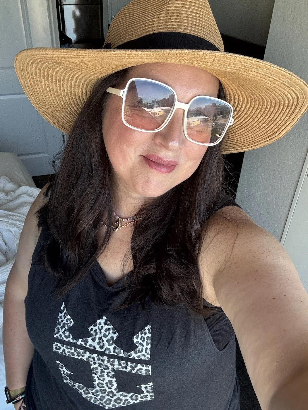 Woman wearing a large straw hat and white sunglasses, taking a selfie indoors. She is dressed in a black tank top with a leopard print design and has a necklace.