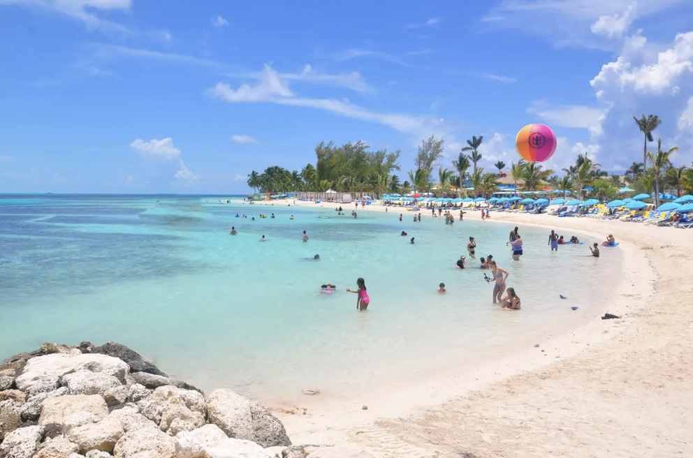 Guests relaxing and swimming along the turquoise shoreline at Chill Island on Perfect Day at CocoCay, with white sand beaches, blue lounge chairs, palm trees, and clear Caribbean water under a sunny tropical sky.