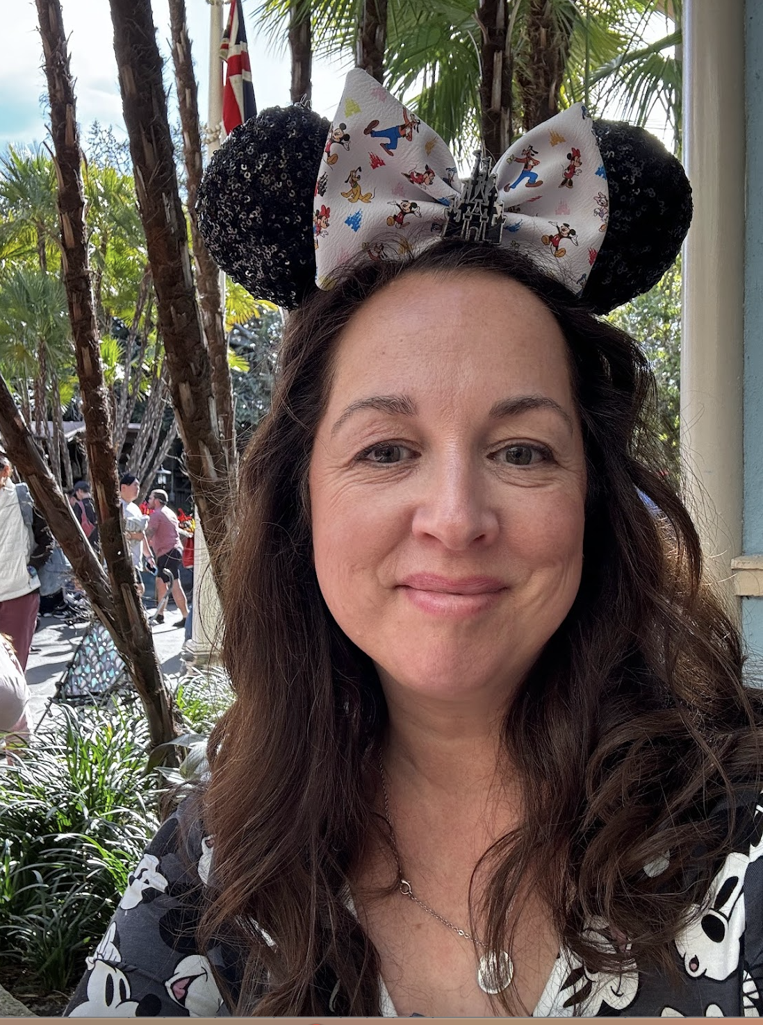A woman with brown, curly hair wearing Mickey Mouse ears and a matching shirt, standing outdoors with palm trees and people in the background.