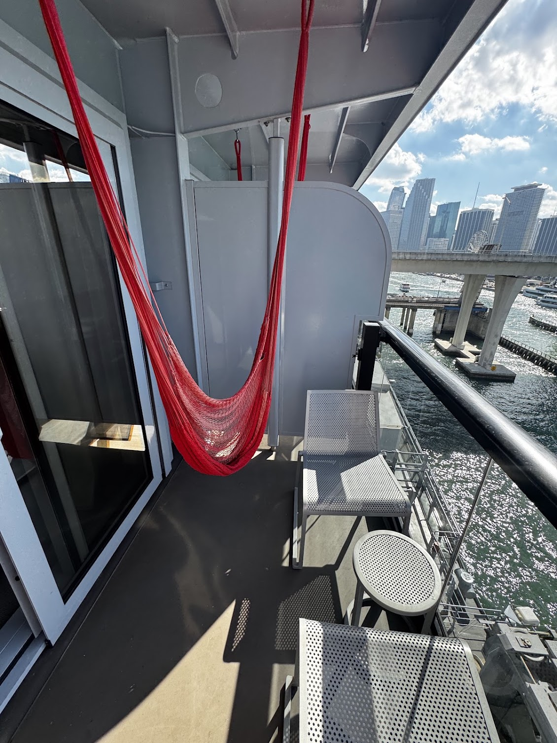 Private balcony on Virgin Voyages cruise with a red hammock, lounge chairs, and views of the Miami skyline from a cruise ship before departure.