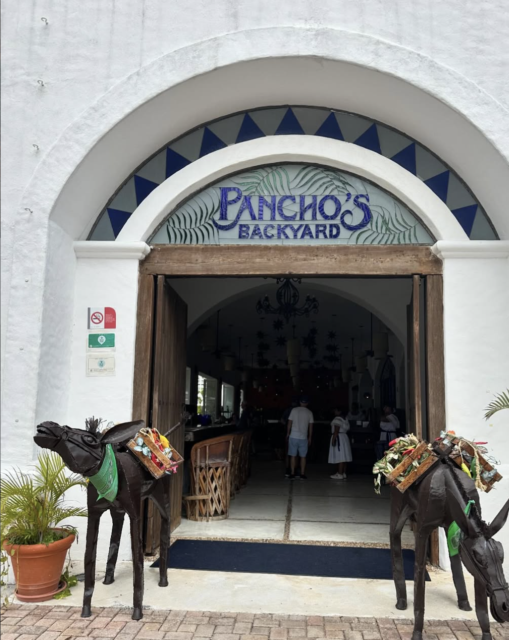 Entrance to Pancho’s Backyard restaurant featuring a white arched doorway with blue stained-glass accents, rustic wooden doors, and decorative donkey sculptures carrying colorful baskets on each side of the entrance.