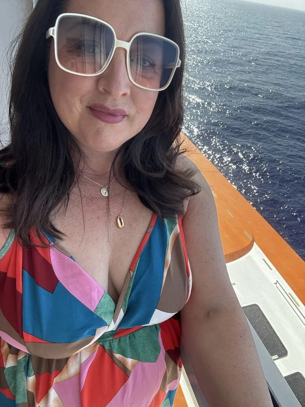 A woman wearing large sunglasses, a colorful sleeveless dress, and layered necklaces takes a selfie on a boat with the ocean in the background.
