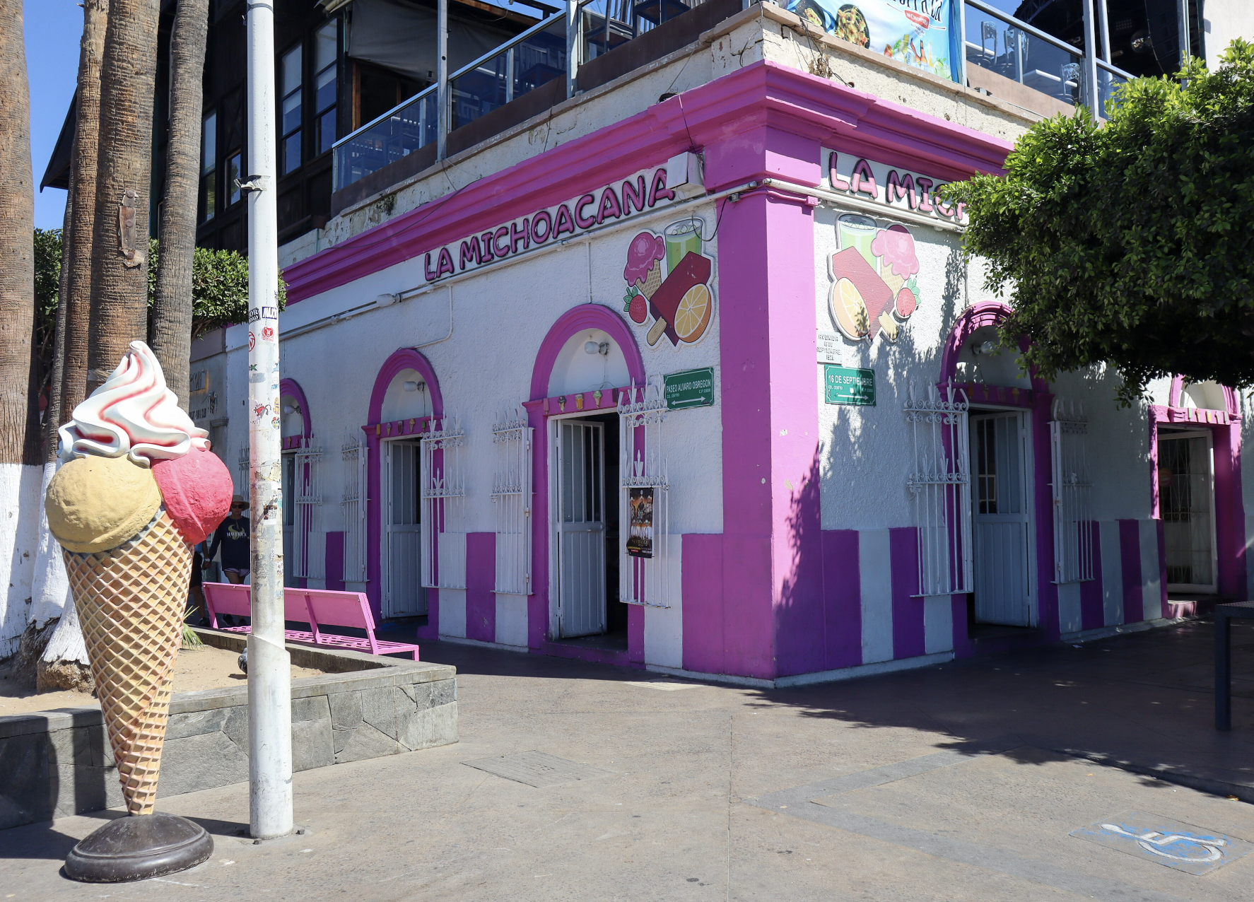 Bright pink and white La Michoacana ice cream shop on a sunny street corner with a giant ice cream cone statue outside and palm trees nearby.