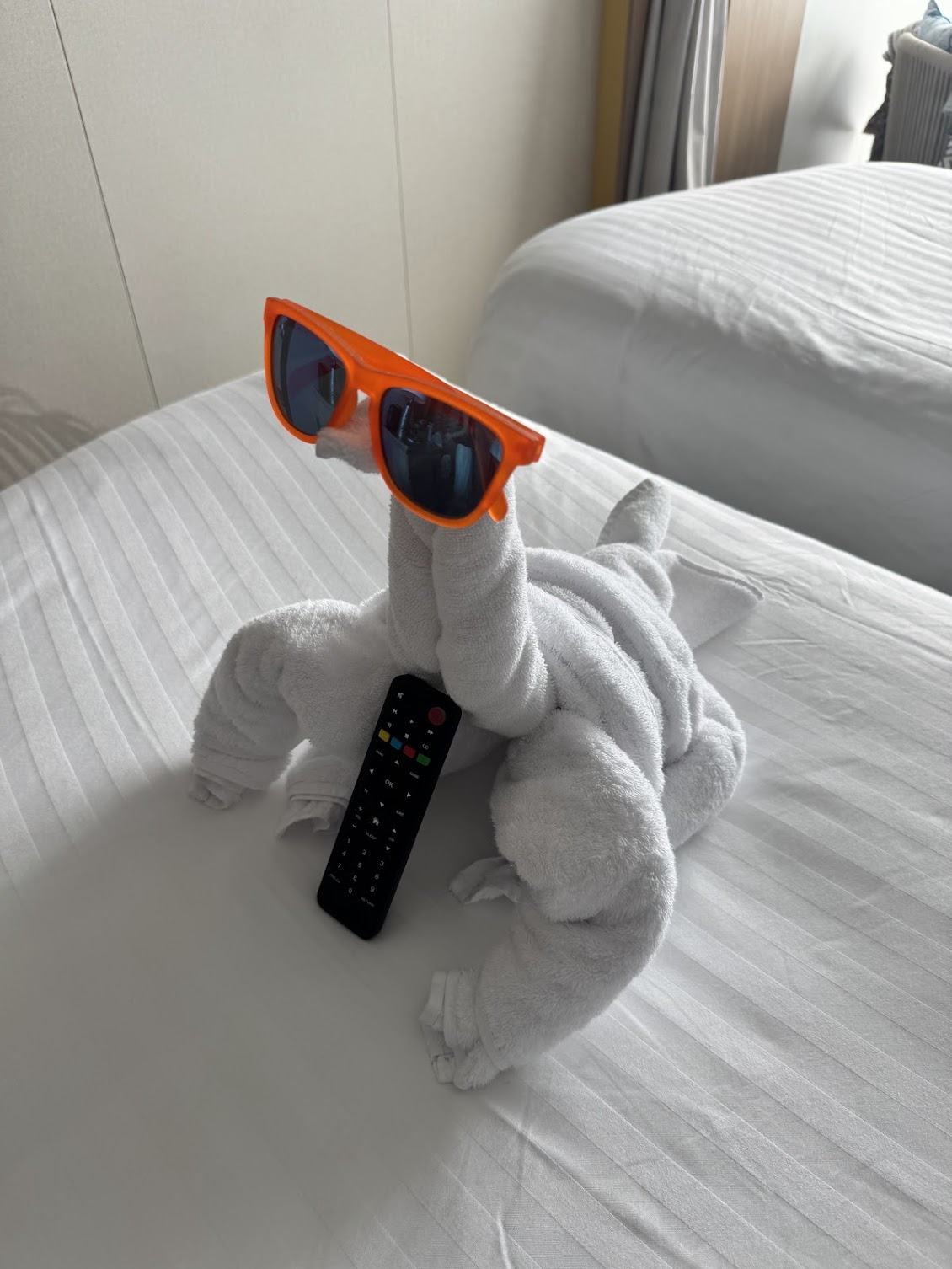 Towel animal wearing sunglasses and holding a TV remote on a freshly made cruise ship bed.