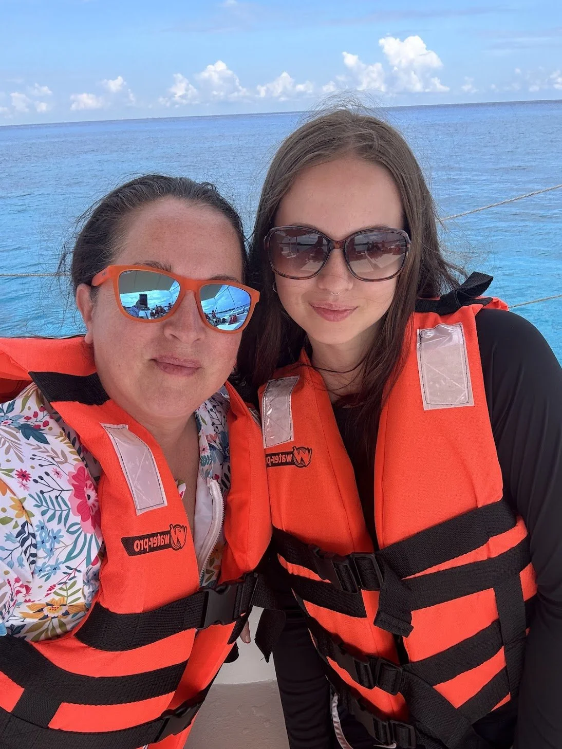 Two women wearing bright orange life jackets and sunglasses posing for a photo on a boat with the turquoise Caribbean Sea in the background.