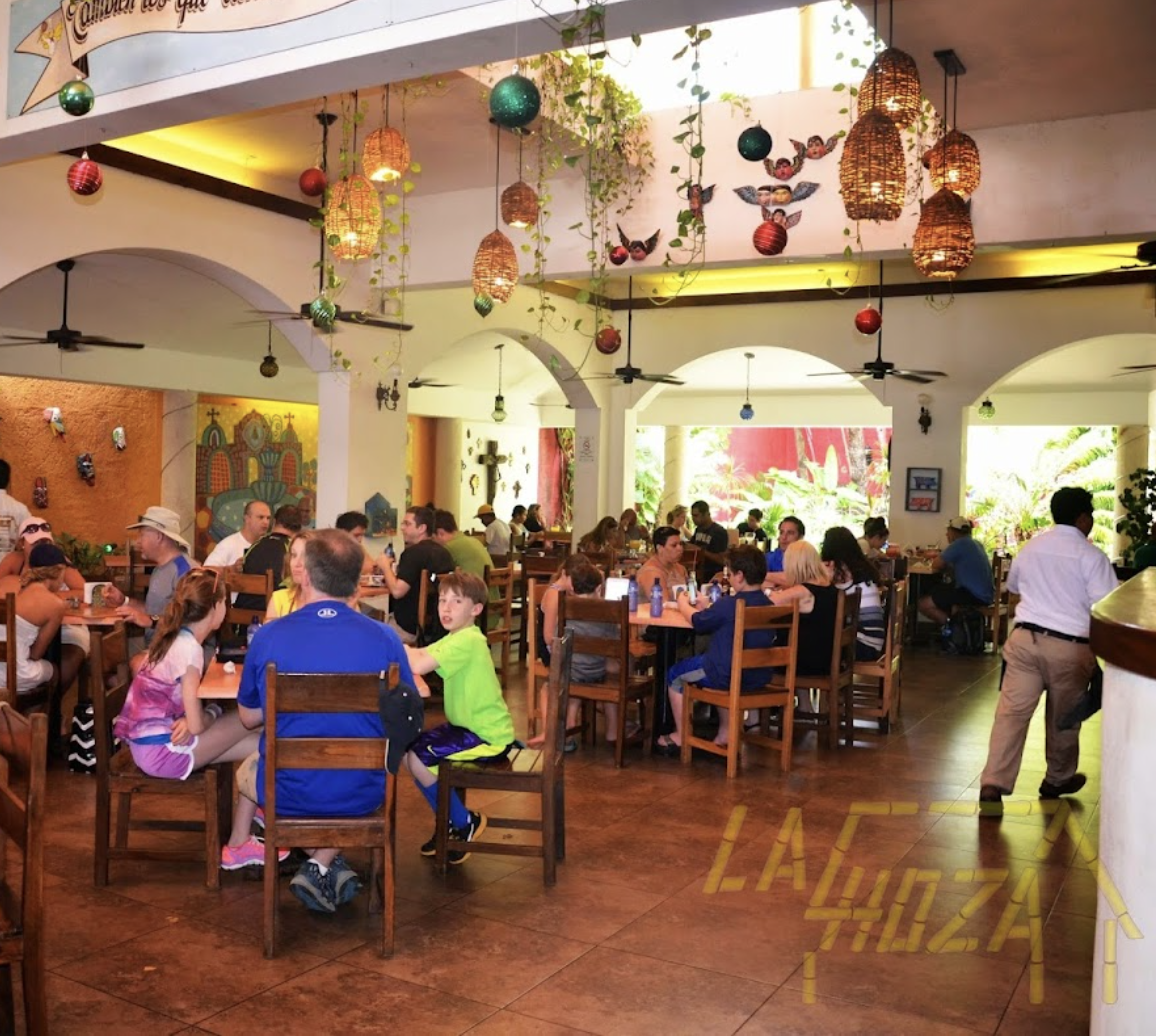 Busy open-air restaurant with colorful hanging lanterns, tropical greenery, and families dining at wooden tables in a warm, vibrant setting.