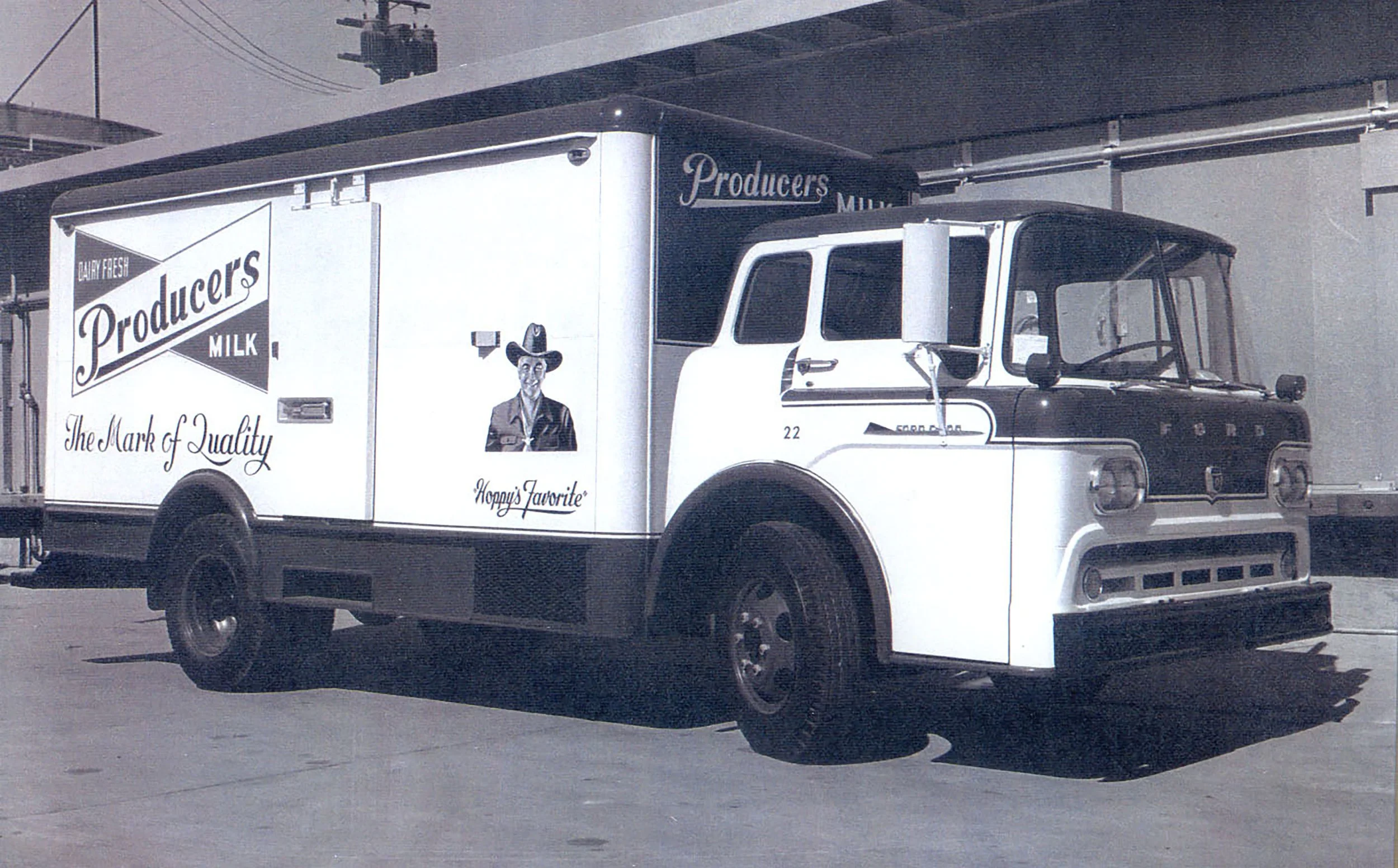 Black and white photograph of a vintage milk delivery truck with advertisements for Producers Milk, including a smiling man in a cowboy hat and the phrases 'The Mark of Quality' and 'Hoppy's Favorite'.