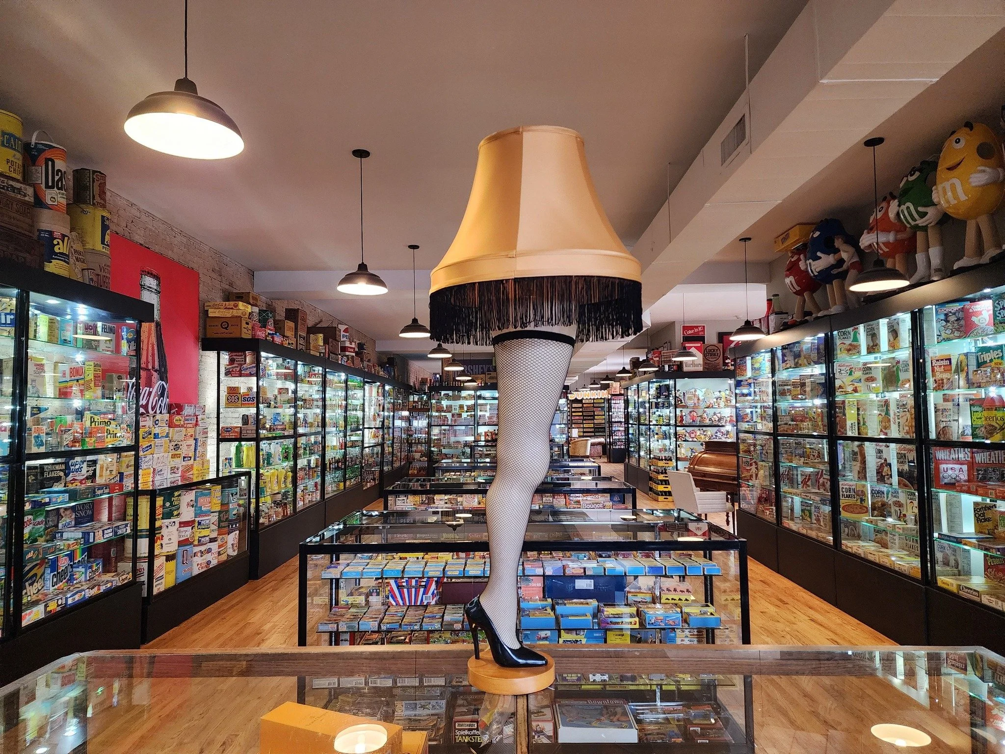 Inside the CBM display cases are filled with various artifacts. A large leg lamp with a fringed lampshade. Shelves on the walls are filled with boxed snacks and toys, including M&Ms and more.