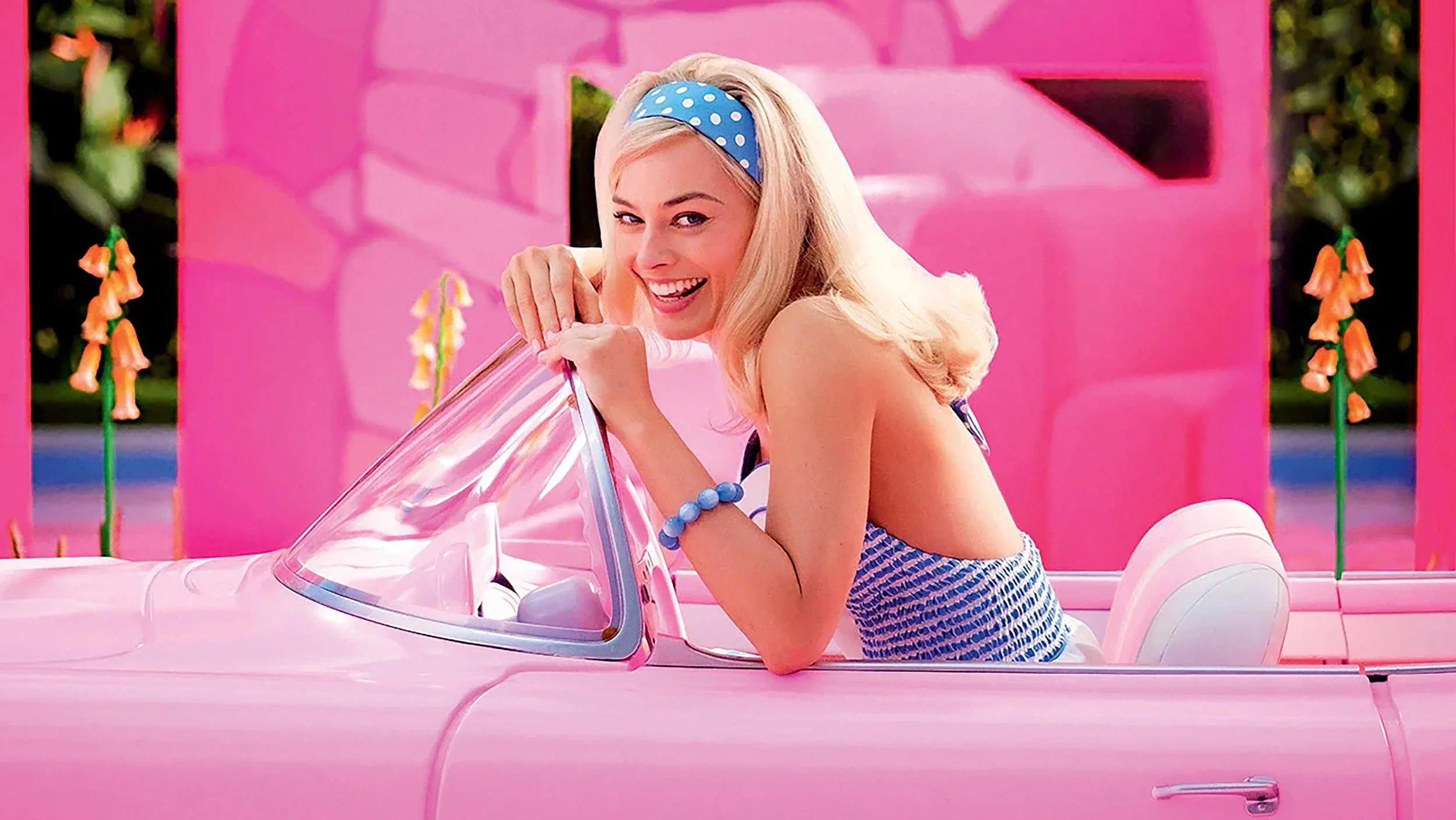 A smiling woman with blonde hair, wearing a blue polka dot headband and blue dress with white stripes, leaning on a pink toy car with a clear windshield, in a pink-themed environment with pink walls and orange flowers.