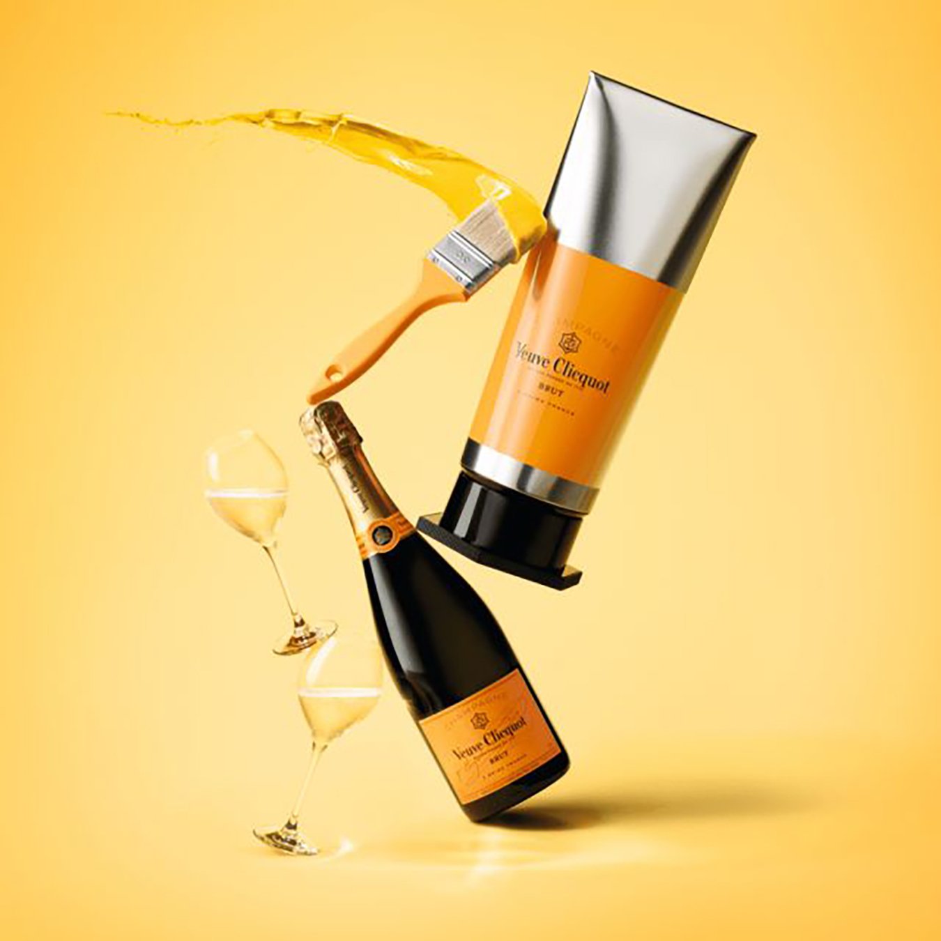 A product shot of a Veuve Clicquot champagne bottle and a tube of skincare cream, with champagne glasses and a champagne bottle in the background on a yellow backdrop.