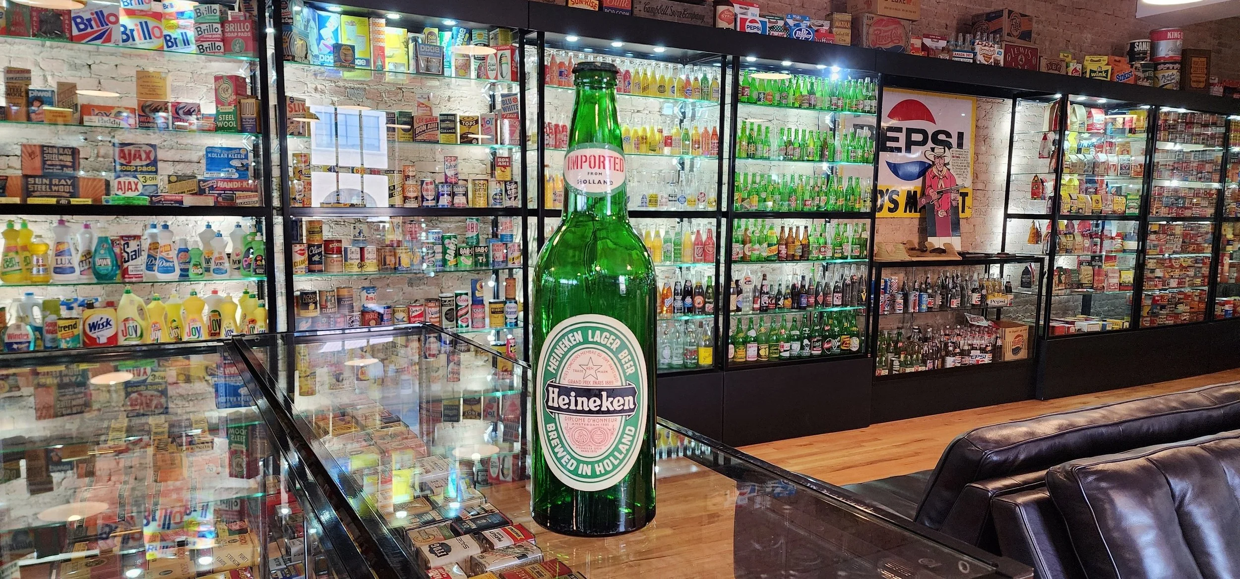 A large Heineken beer bottle with a green label inside the CBM. The background features shelves filled with various colorful products, including bottles, cans, and boxes, along with an old brick wall and a Pepsi sign.