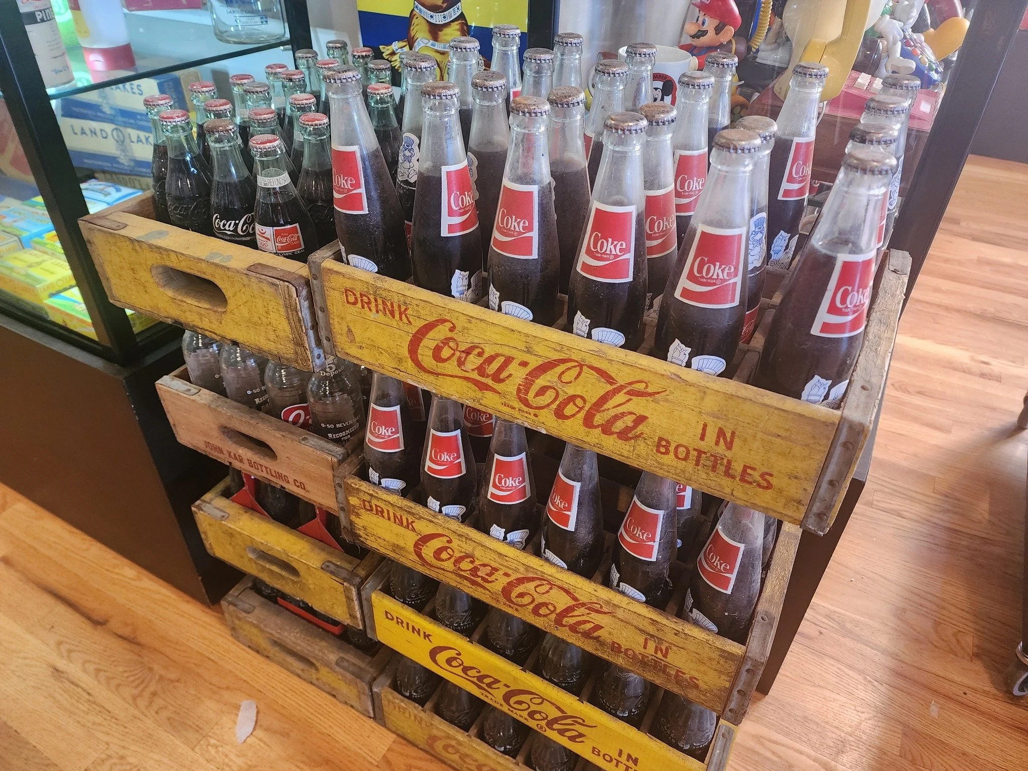 A wooden display stand filled with glass bottles of Coca-Cola in a store. The bottles have vintage-style labels and are arranged upright. The stand has yellow and red branding and the word 'Drink' and 'In Bottles' written on it. In the background, pa