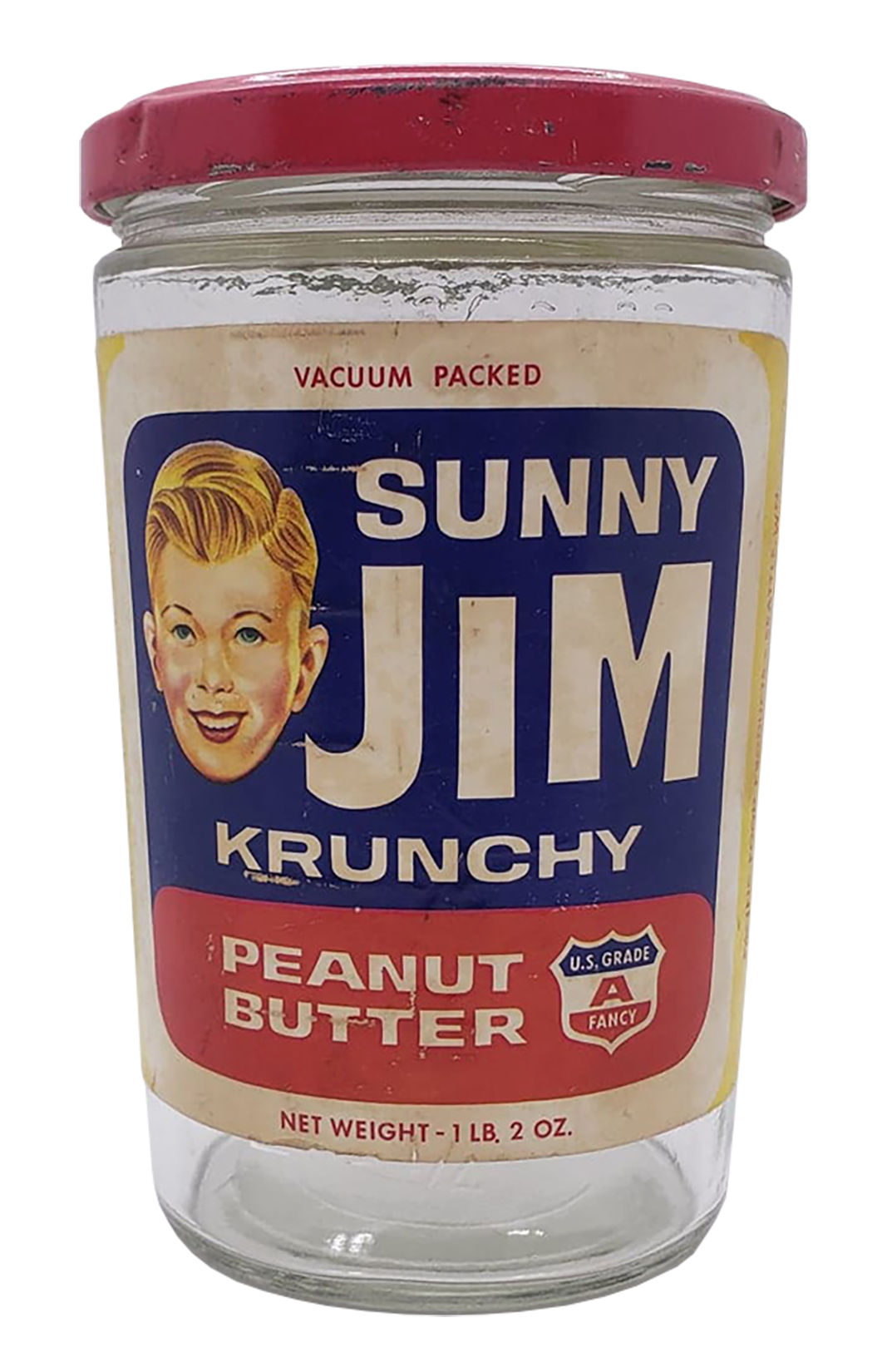 A glass jar of Sunny Jim Krunchy Peanut Butter with a red screw-on lid and vintage label, showing a smiling boy's face and text indicating it's vacuum packed, 1.2 ounces weight, and U.S. grade A fancy quality.