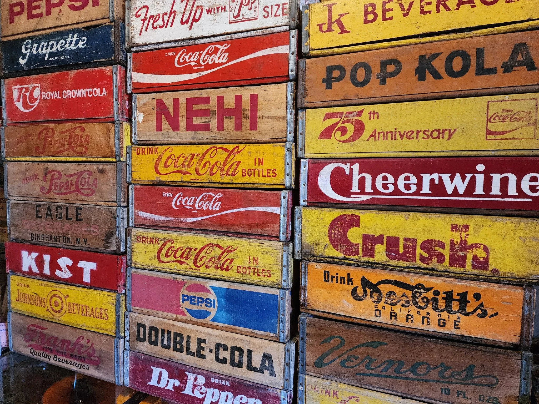 Stacked vintage wooden crates advertising various soft drinks and beverages, including Coca-Cola, Pepsi, Georgia Peach, Ginger Beer, and others, with colorful and retro designs.