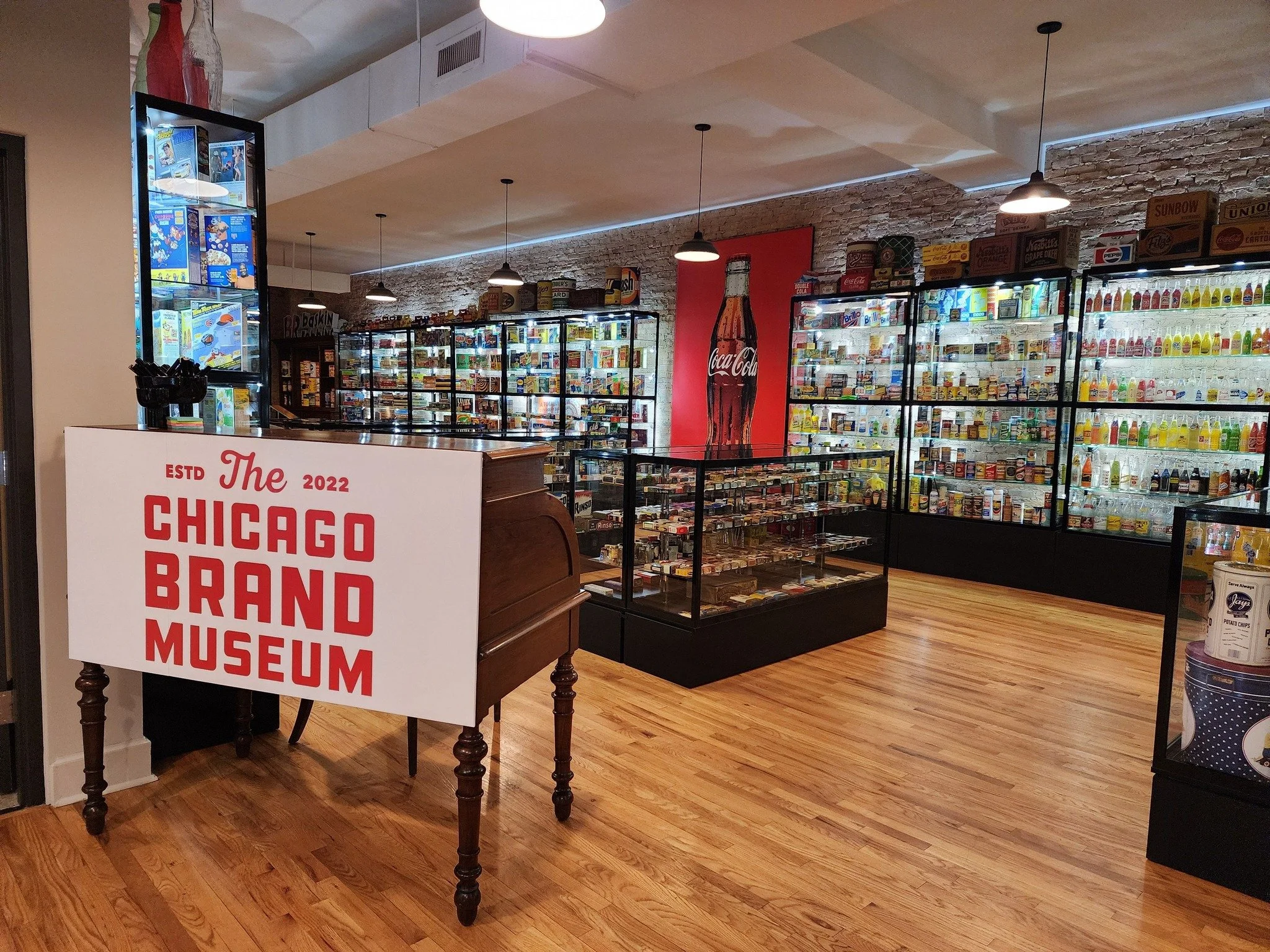 Interior of the CBM display cases filled with artifacts, brick walls, wooden floors, and ceiling lights. A sign on a wooden table reads 'The Chicago Brand Museum,' and there is a large Coca-Cola advertisement on the brick wall.