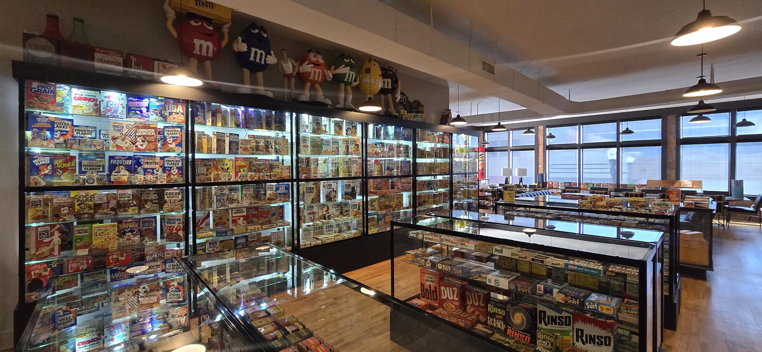 Interior of the museum. Cabinets are filled with vintage cereal boxes and artifacts. There are large M&M's displays on a shelf above the cabinets. The room has large windows with city view and industrial-style ceiling lights.