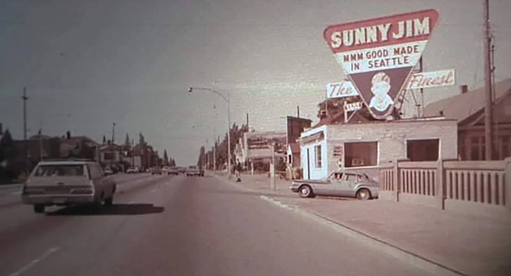 A street scene with parked cars, street lamps, and a large vintage sign on a building that reads 'Sunny Jim' and features a cartoon character, with other buildings and trees in the background.