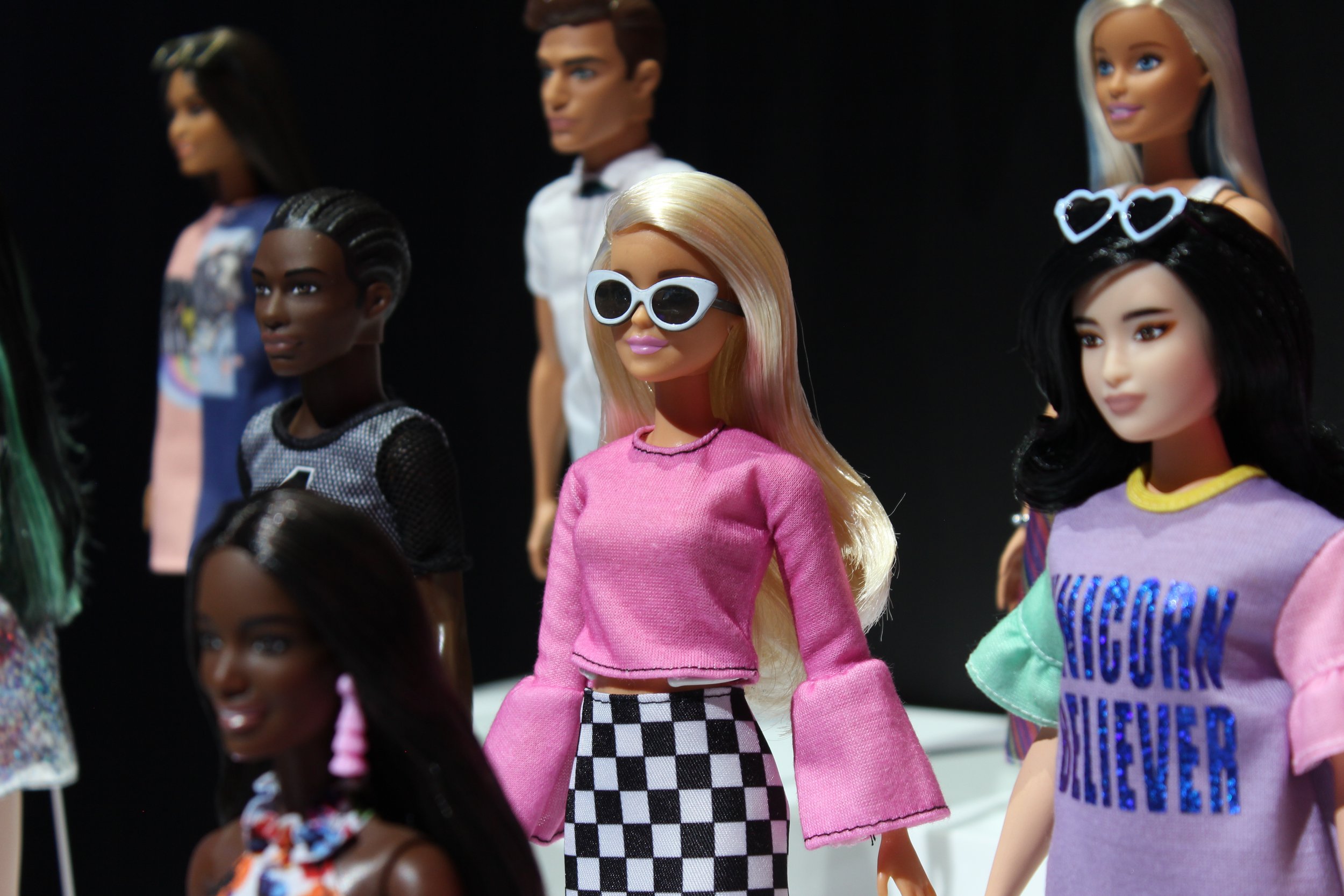 Group of diverse fashion dolls dressed in trendy clothing, with some wearing sunglasses and colorful outfits, set against a black background.