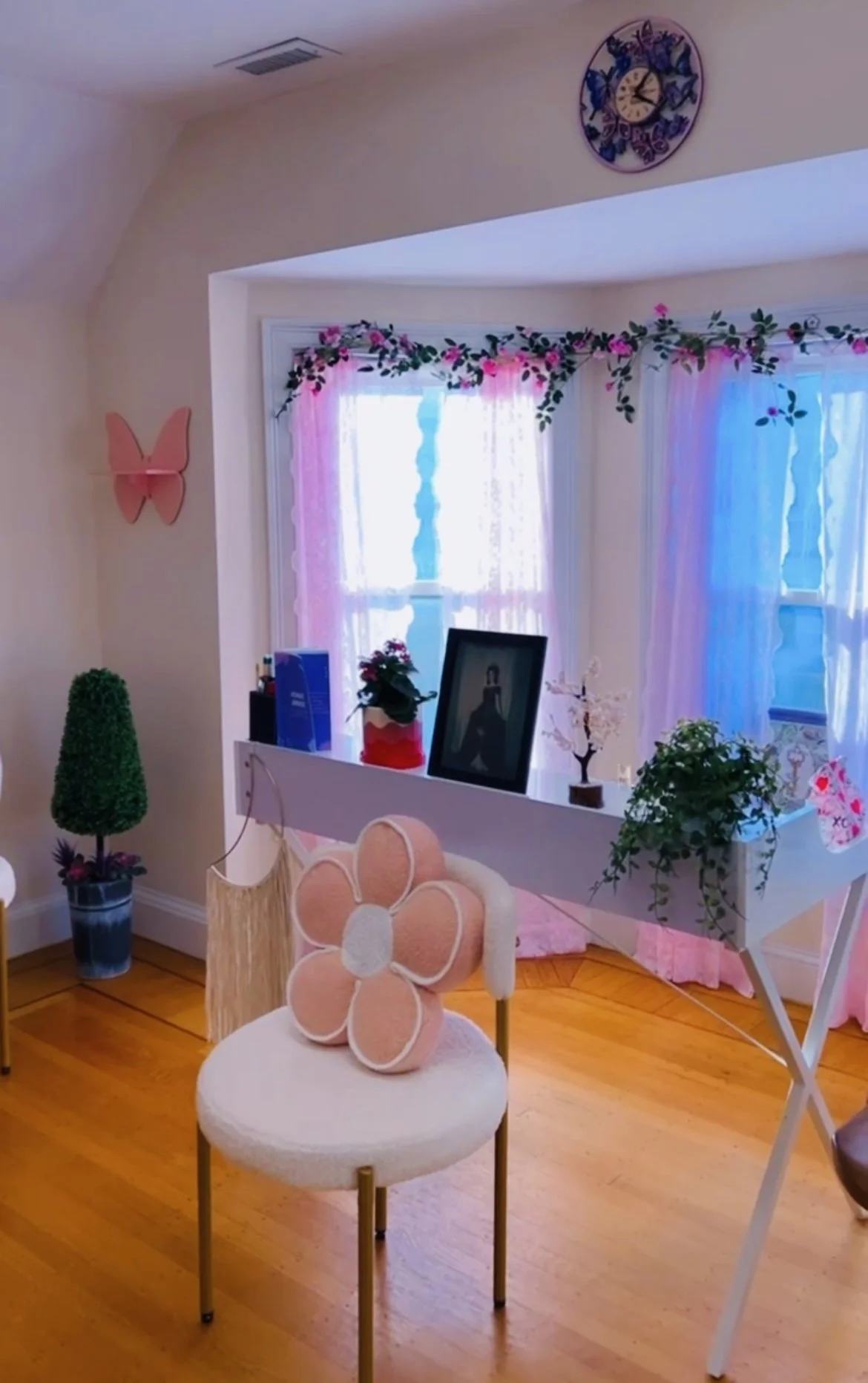 Decorated cozy corner with pink, blue, and white accents, a small chair with a flower pillow, plants, framed photo, and pink curtains with floral garland.