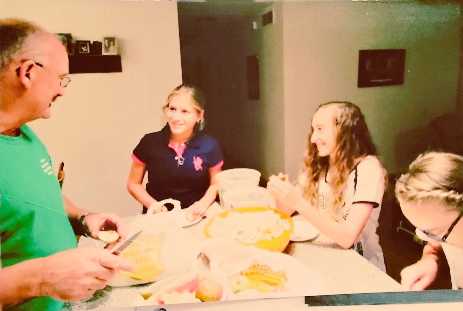 A group of people gathered around a kitchen table, preparing food. The man is slicing something while others are interacting and smiling. Various food items are visible on the table.