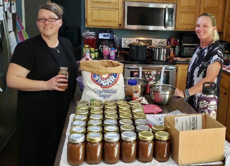 Two people in a kitchen with a table full of jars, granulated sugar bag, and kitchen utensils.