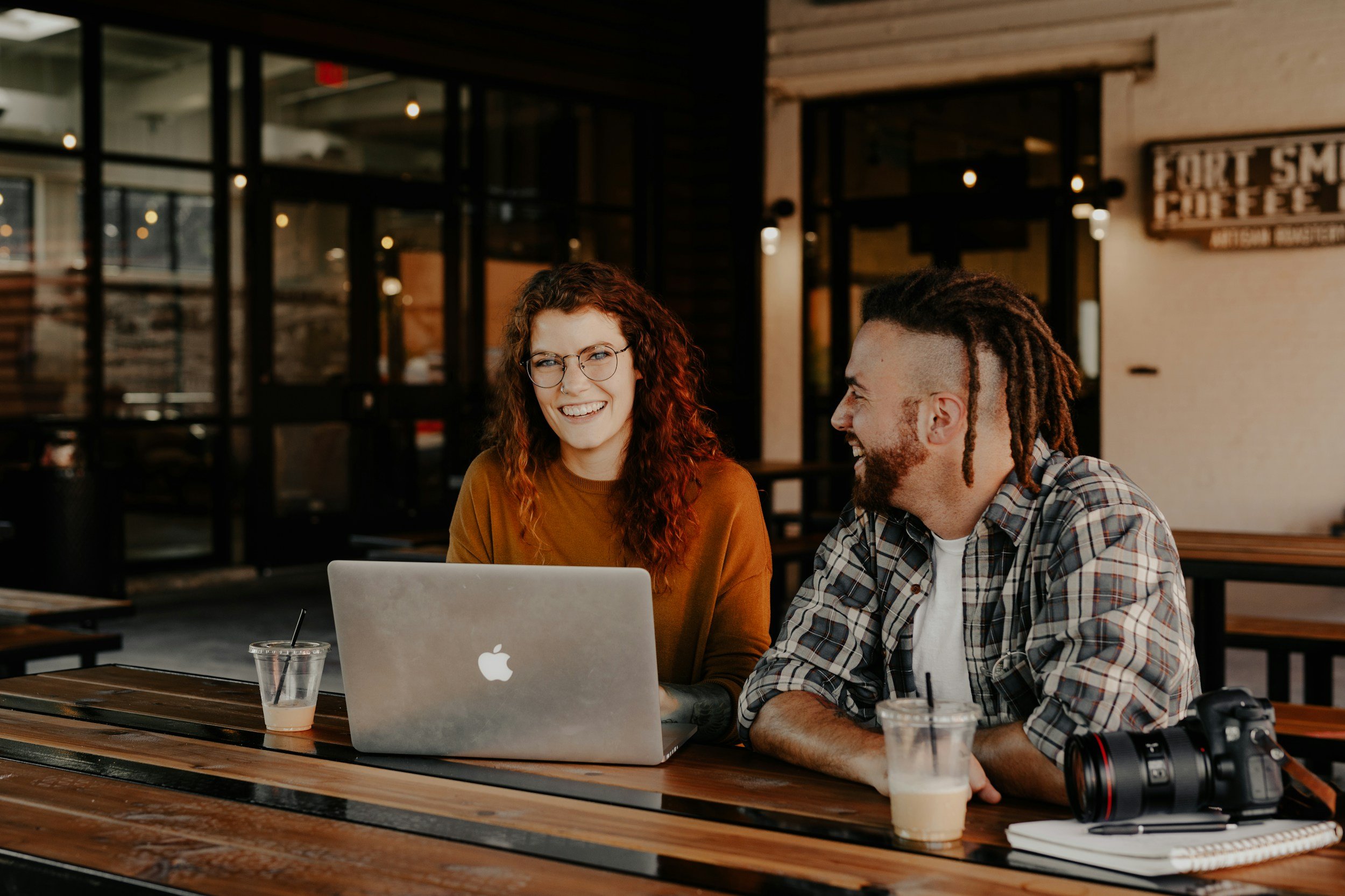 Two people sitting at an outdoor café table, smiling and chatting while working for a nonprofit on a laptop.
