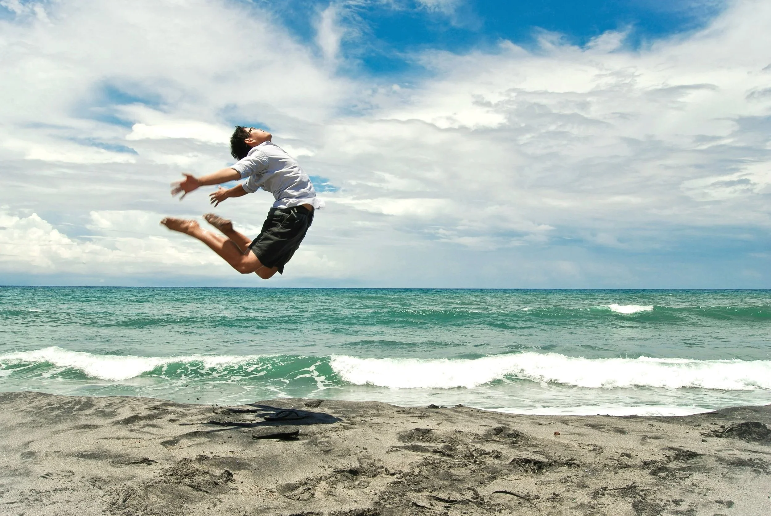 A person jumping on the sandy beach with the ocean and cloudy sky in the background.