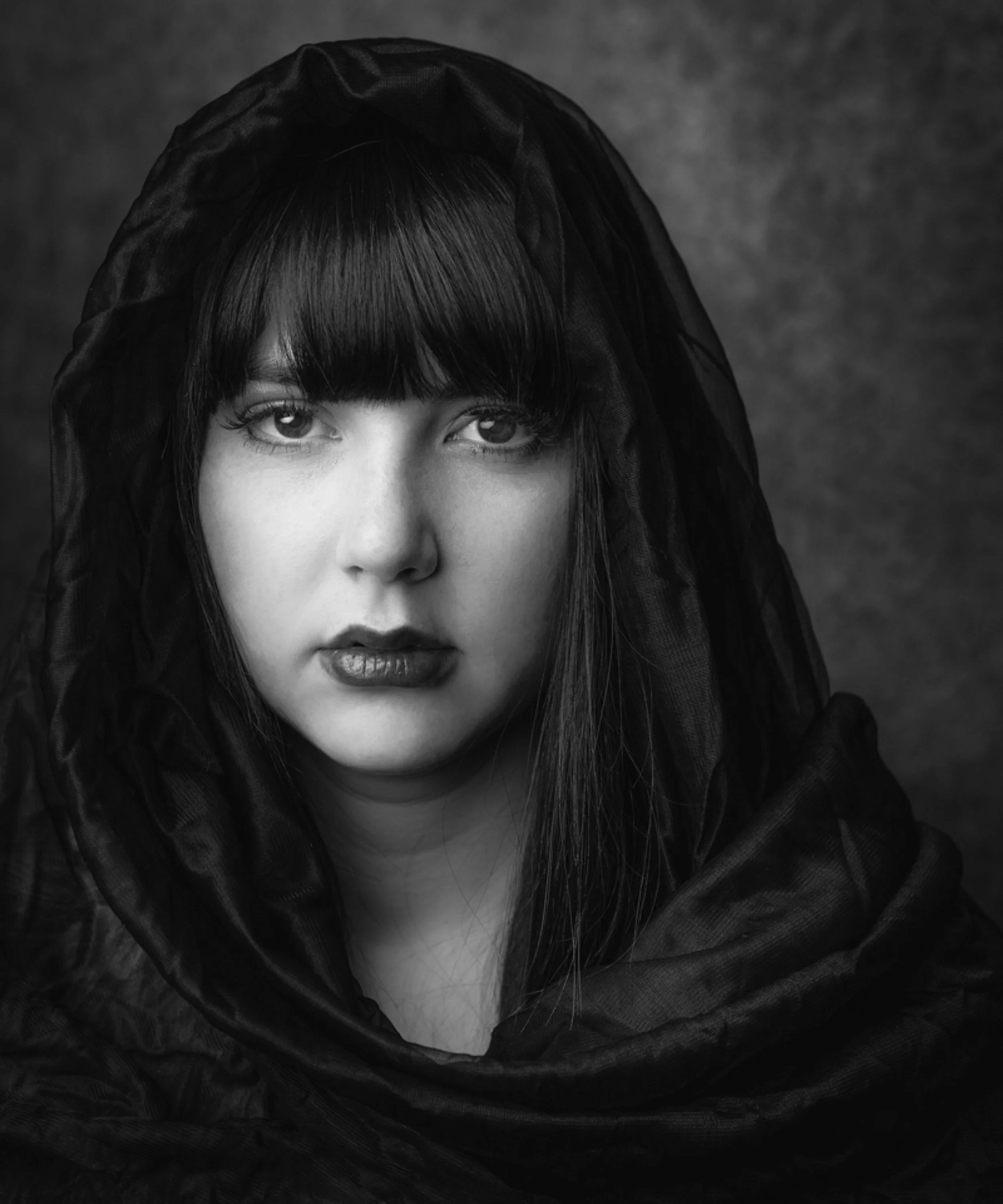 Close-up black and white portrait of a woman with dark hair and bangs, partially covered with a black fabric or veil, against a dark background.