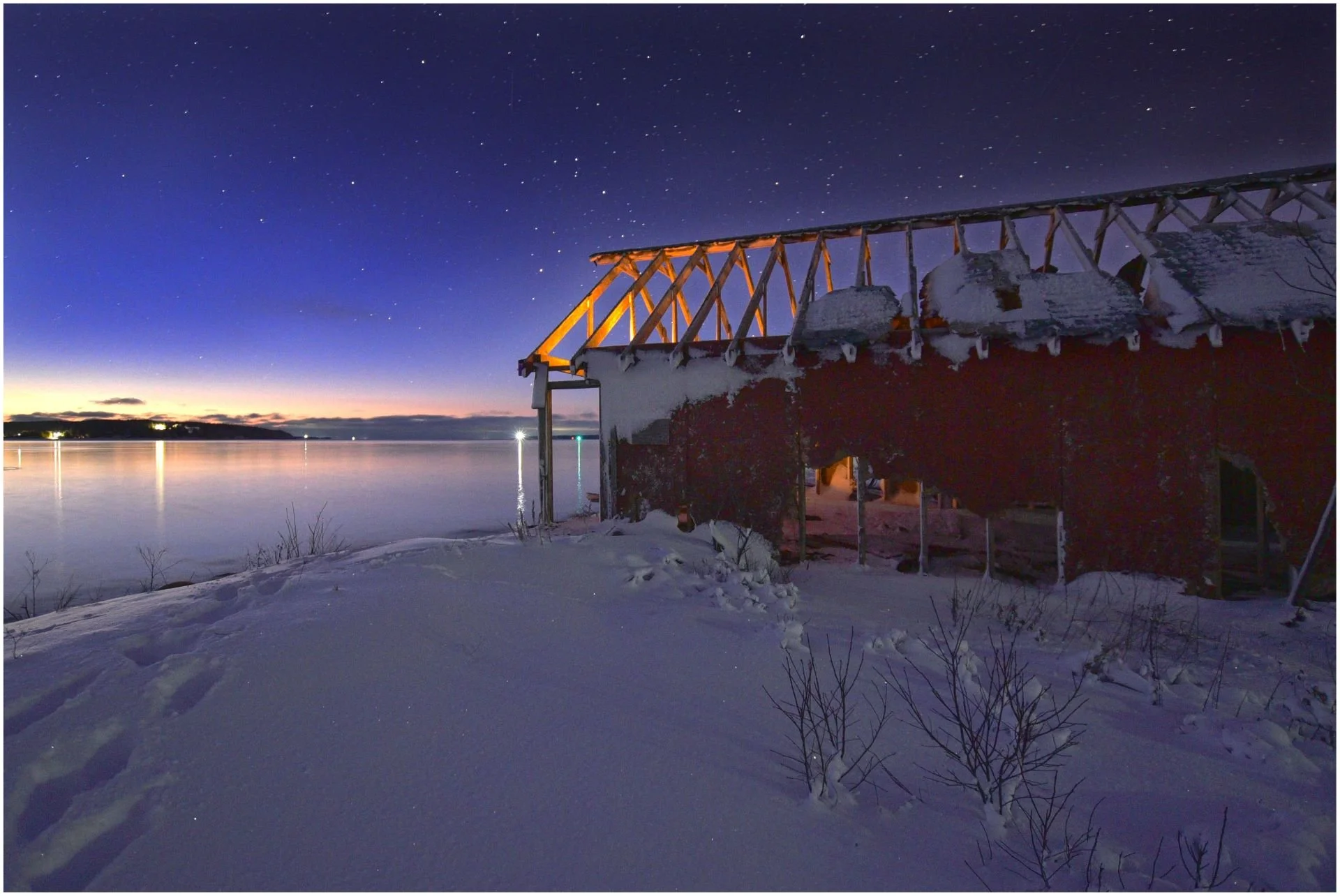 Snow-covered shoreline with an abandoned building in the foreground. The sky is clear with numerous stars visible, and the water reflects the twilight colors.