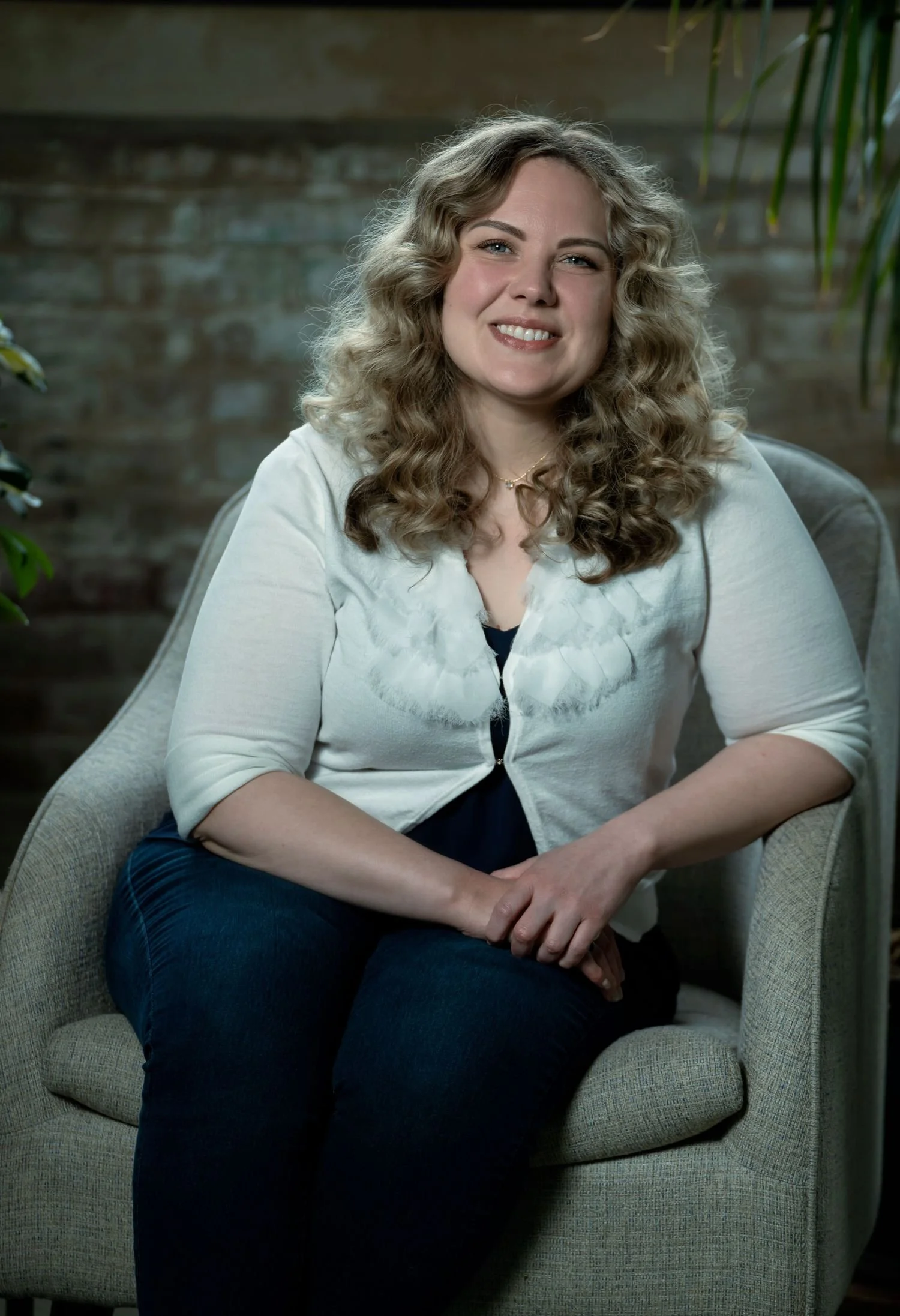 A woman with curly blonde hair and light skin is sitting in a beige armchair in front of a brick wall. She is smiling and wearing a white cardigan over a dark top and dark pants.